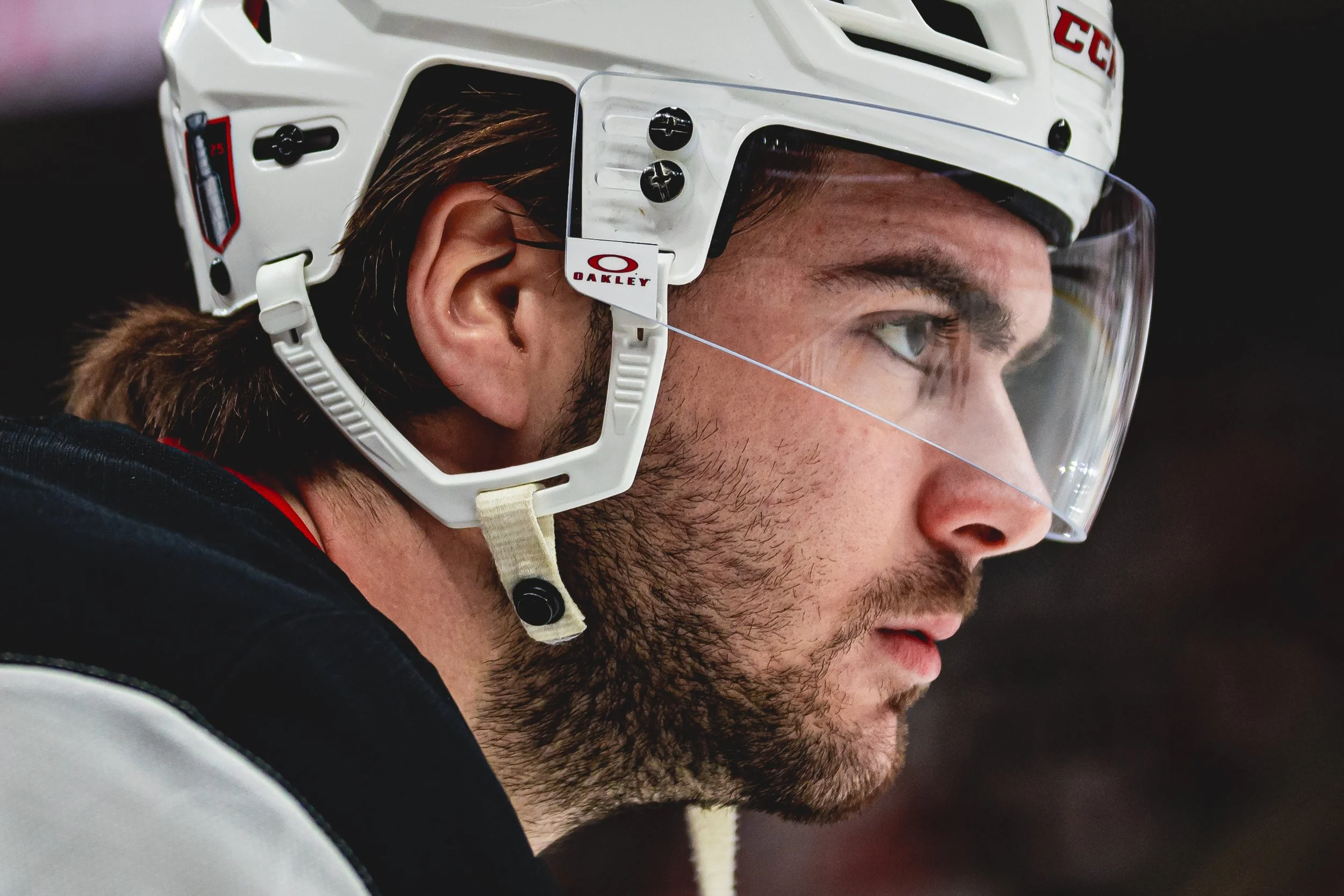Close-up of a male ice hockey player wearing a white helmet with safety visor, focusing intently, with brown hair and a beard visible.