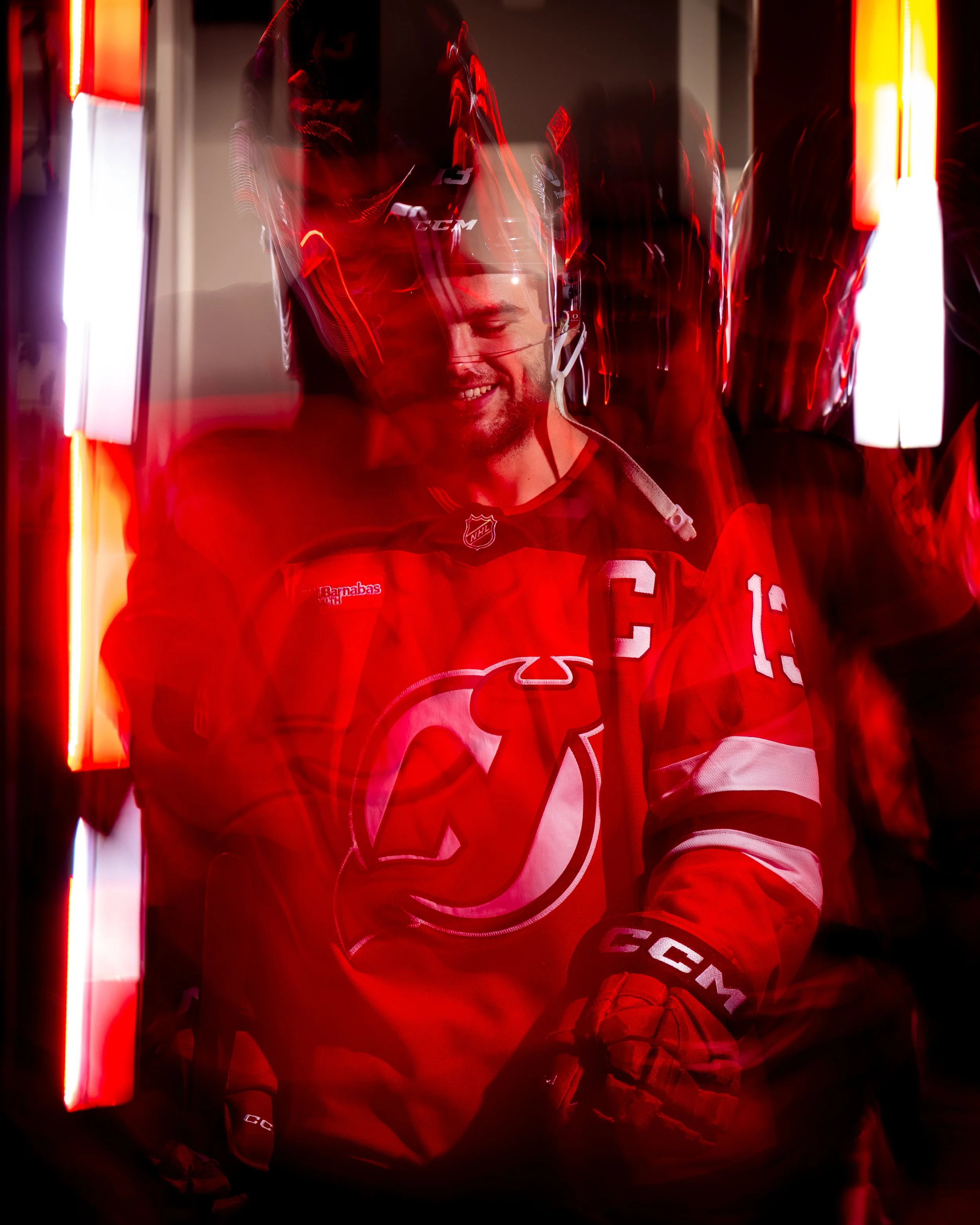 A smiling hockey player wears a red jersey with the New Jersey Devils logo, a helmet, and gloves, standing in front of a locker room with red vertical lights.