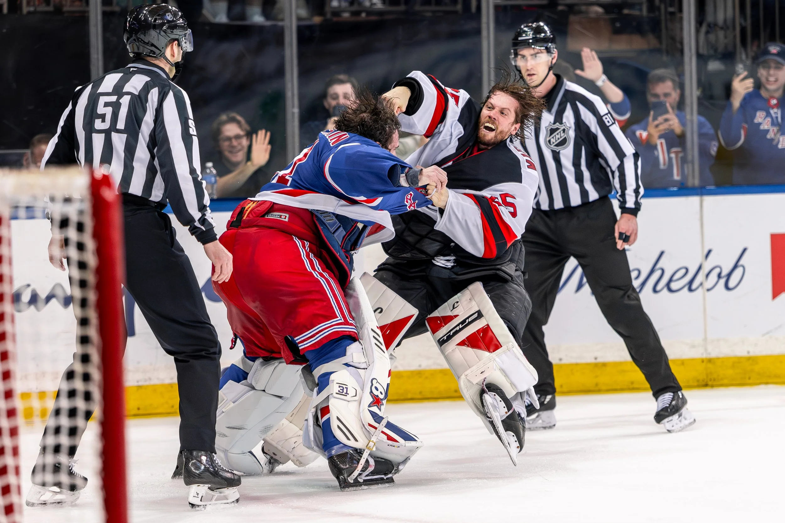 A hockey fight breaking out on the ice between players, with referees attempting to intervene, while spectators watch and record the scene from behind the glass.
