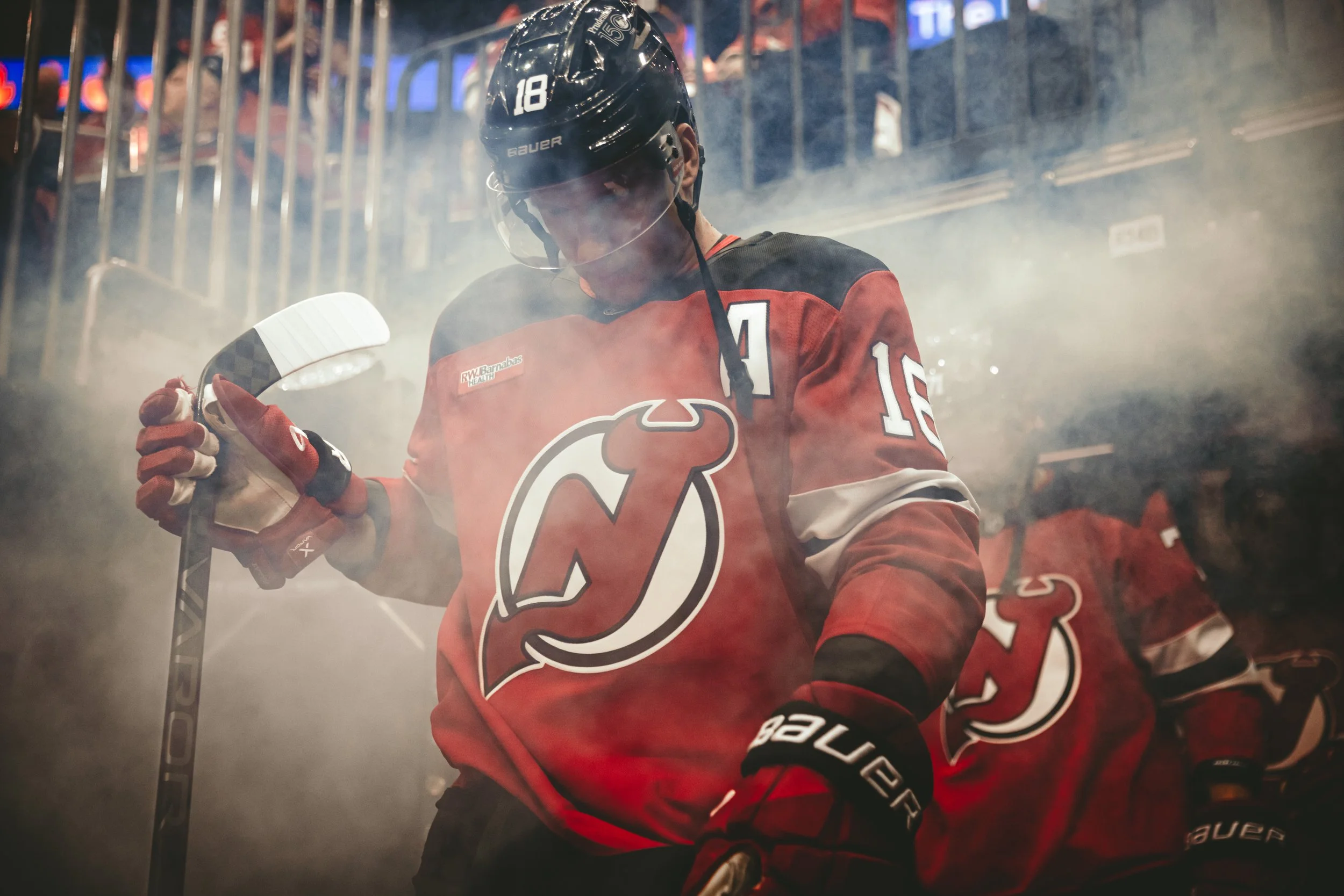 An ice hockey player in a red and black jersey with the number 18 and a team logo, holding a hockey stick, walking through smoke or fog in an arena.
