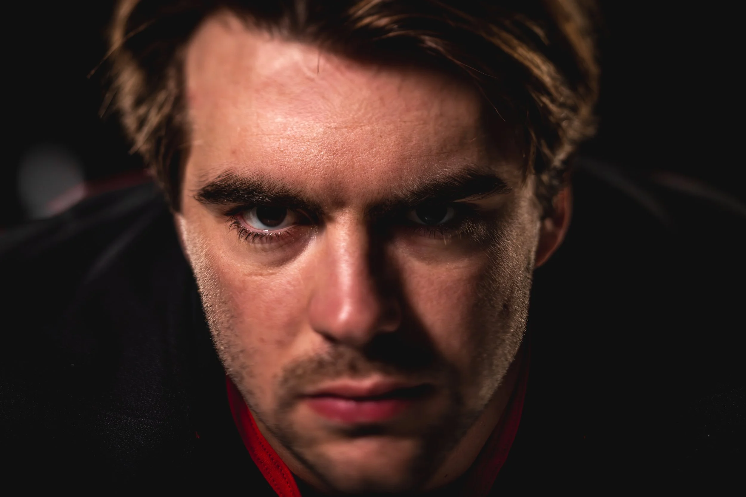 A close-up of a young man with intense eyes and brown hair, wearing a black hoodie with red trim, against a dark background.