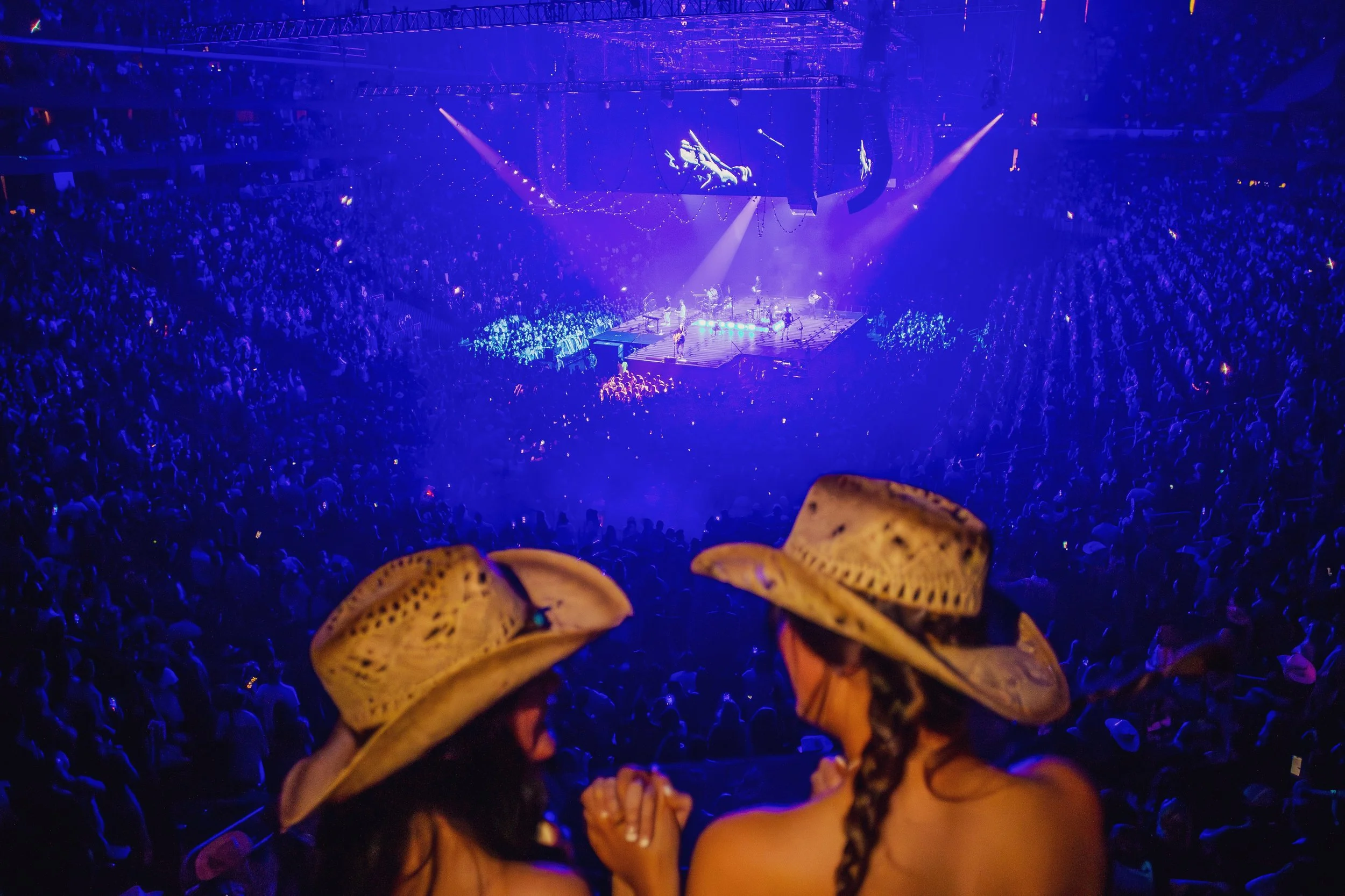 Concert inside a large arena with a stage illuminated by purple lights, a big crowd seated and standing around, and a tall screen displaying performers.