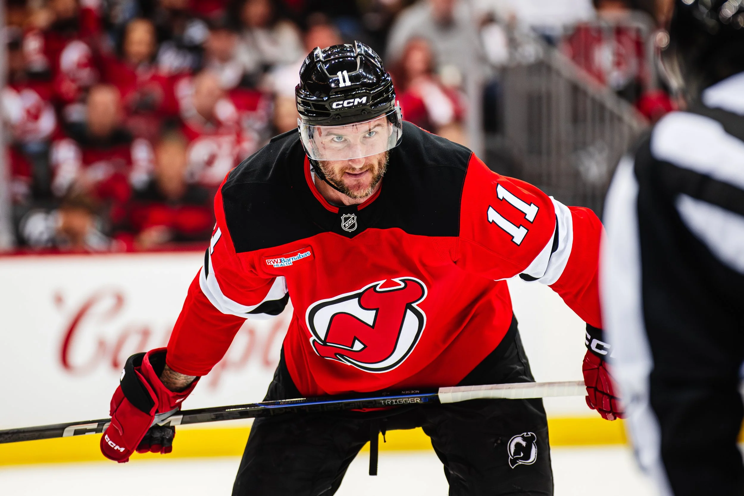 A hockey player wearing a black helmet and red jersey with the number 11 on it, playing on the ice during a game, with a crowd of spectators blurred in the background.