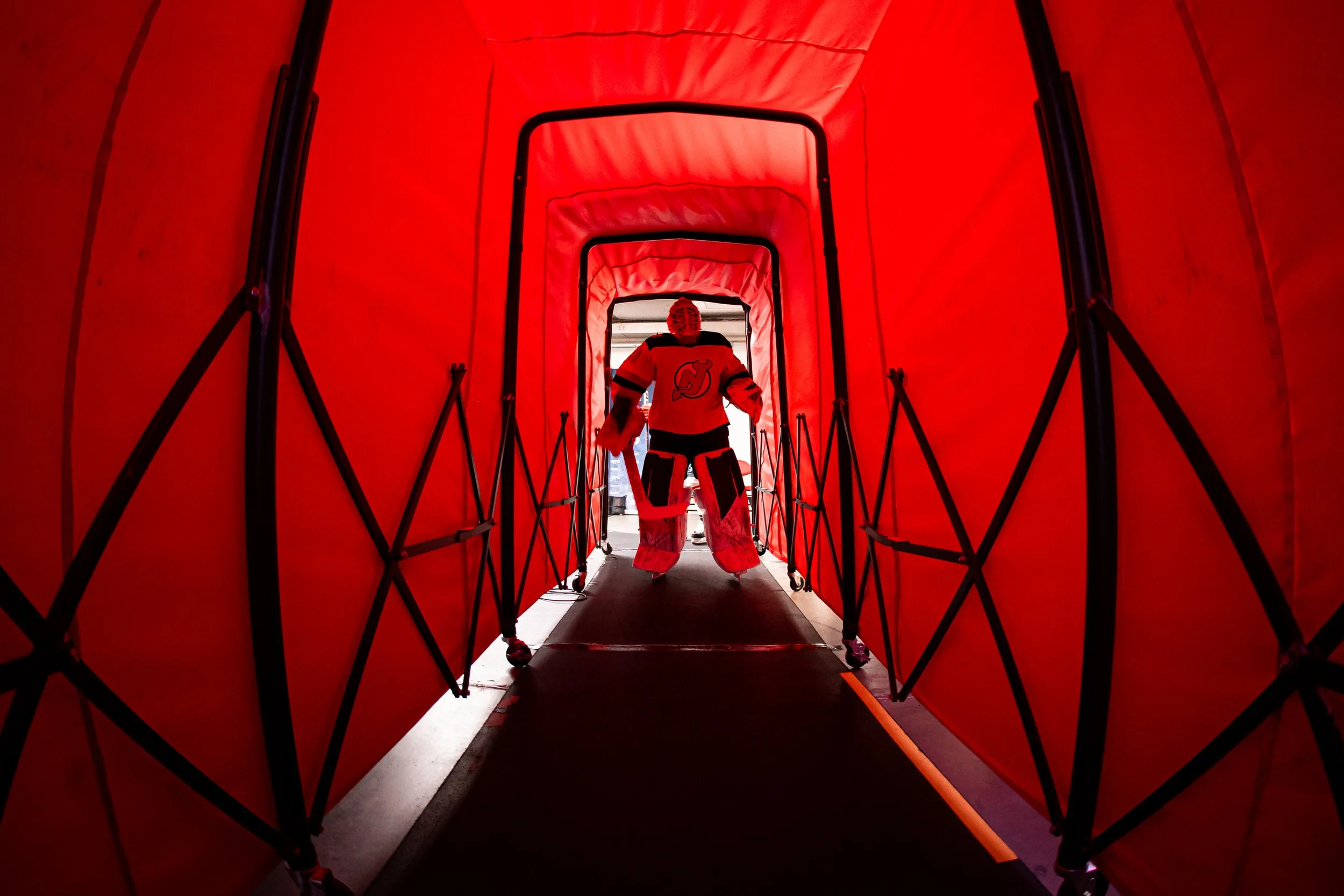 Hockey goalie walking through a red tunnel towards the ice rink, wearing a helmet and team uniform.
