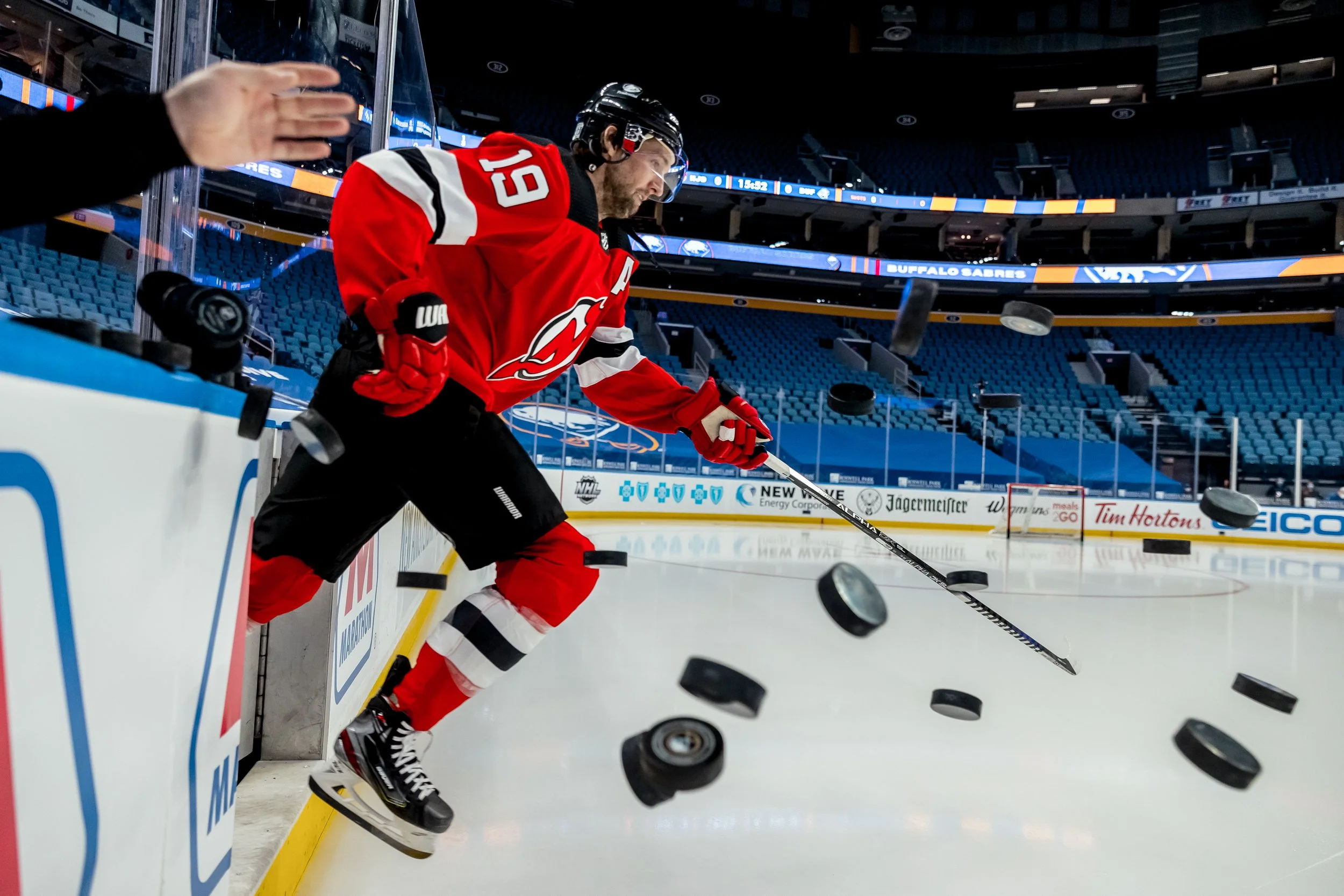 An ice hockey player in red uniform with the number 19, wearing a helmet and gloves, is practicing near the bench as pucks scatter on the ice during a game or practice in an empty arena.