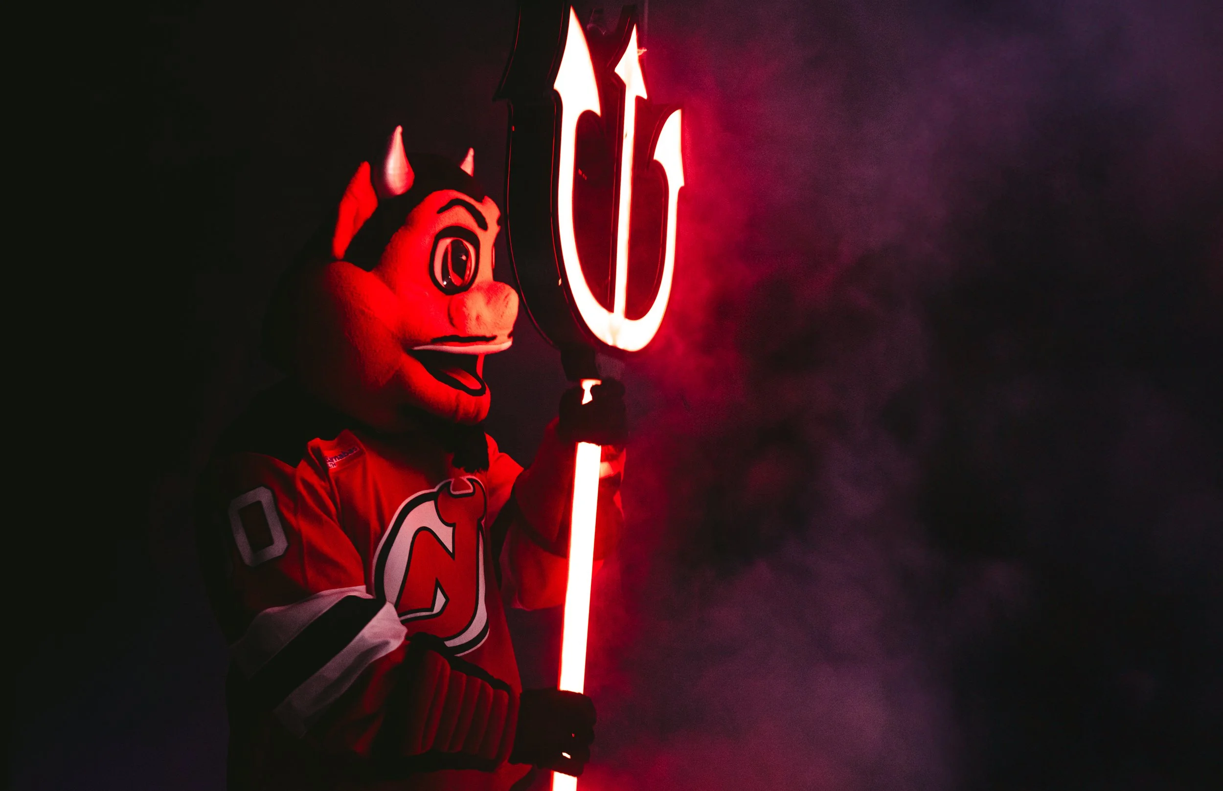 A person dressed in a New Jersey Devils hockey jersey and a devil mascot hat, holding a glowing red trident with a neon Devils logo glowing behind them in a smoky, dark setting.
