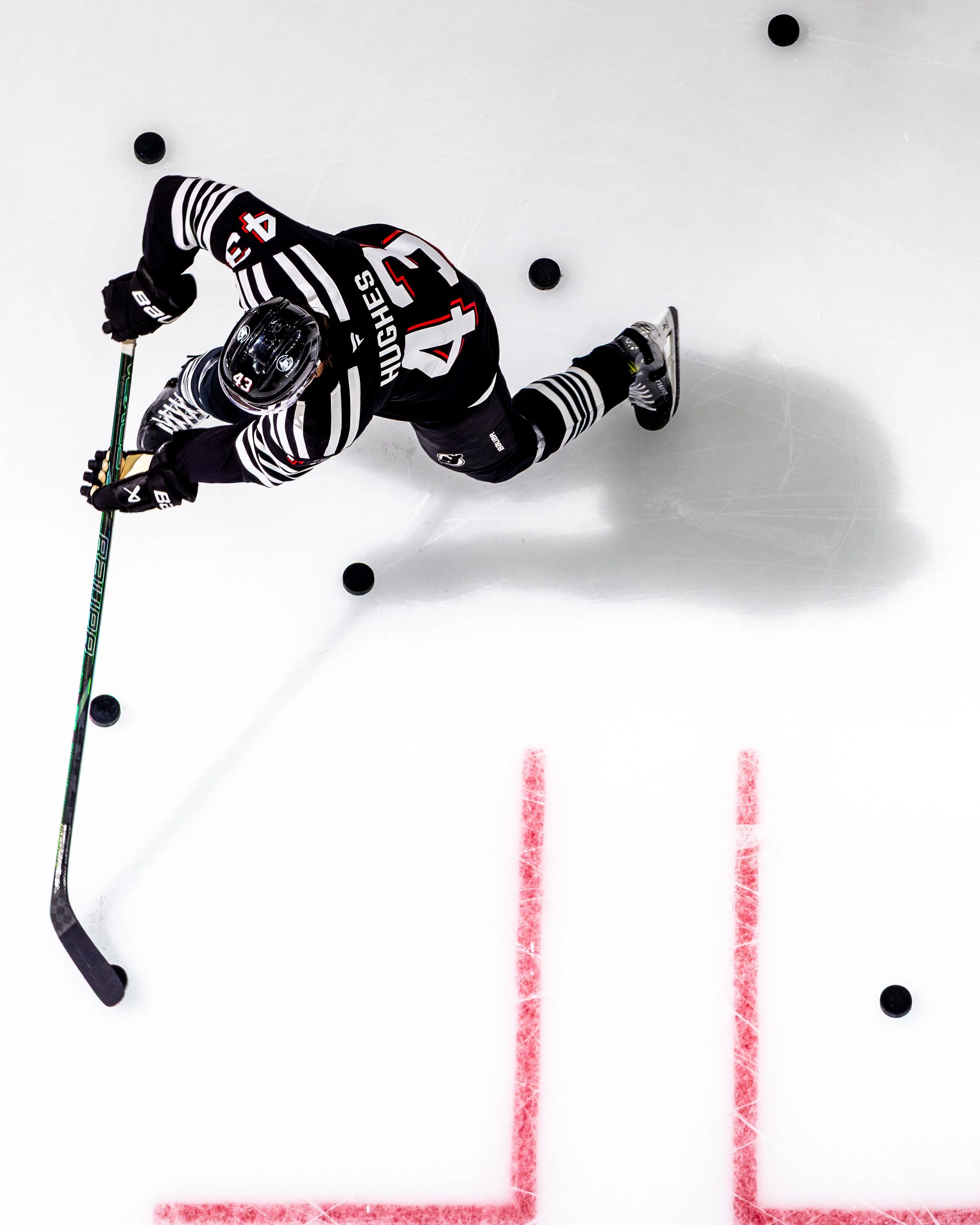 An overhead view of an ice hockey player in a black jersey with red and white accents, wearing a helmet and skates, crouching on the ice near two red goal lines with hockey pucks nearby.