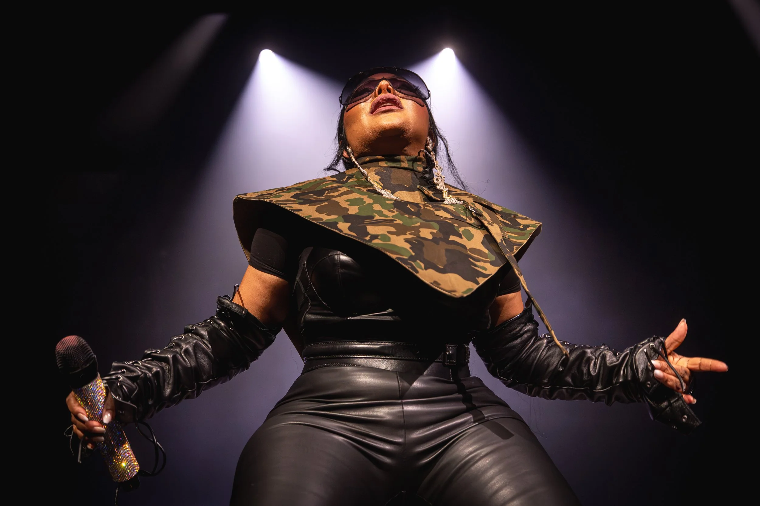 Female performer on stage wearing sunglasses, camouflage top, and black leather pants, with lighting and microphone in hand