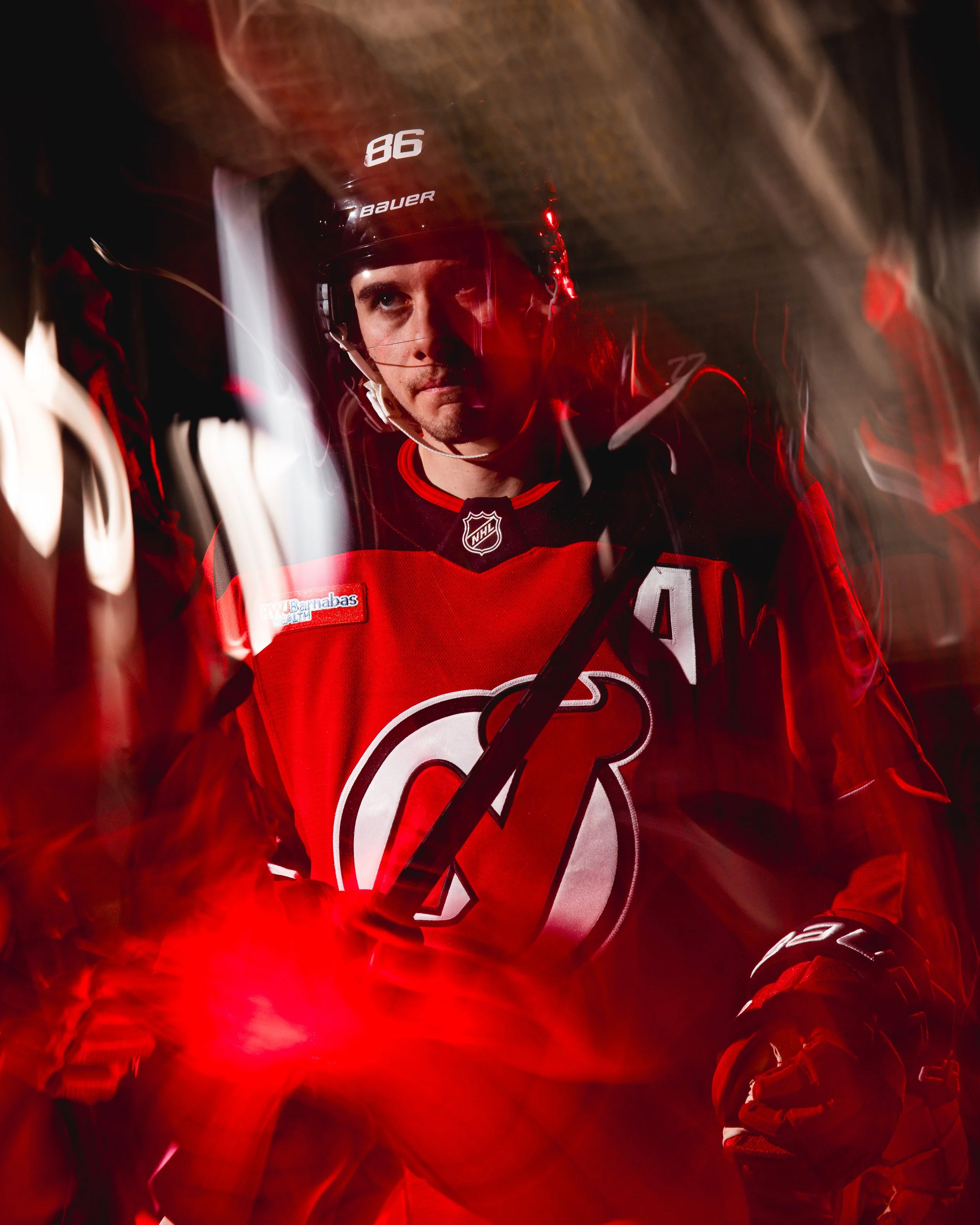 A male ice hockey player in a red jersey and black helmet with the number 86, looking sideways with a serious expression in a dynamic, motion-blurred scene.