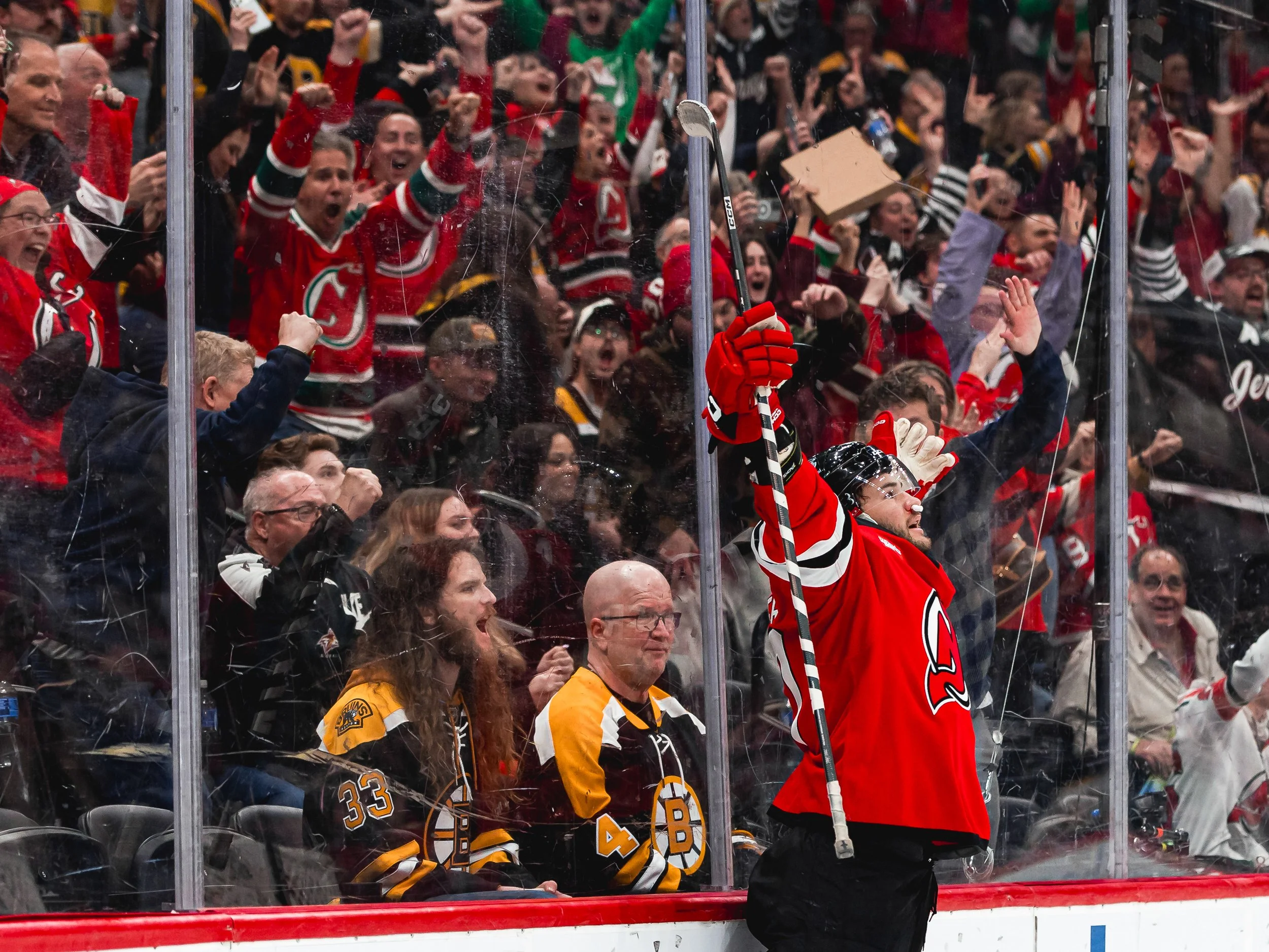 A large group of hockey fans in team jerseys celebrating behind a glass barrier at a hockey game, with one player in a red jersey raising his stick and cheering.
