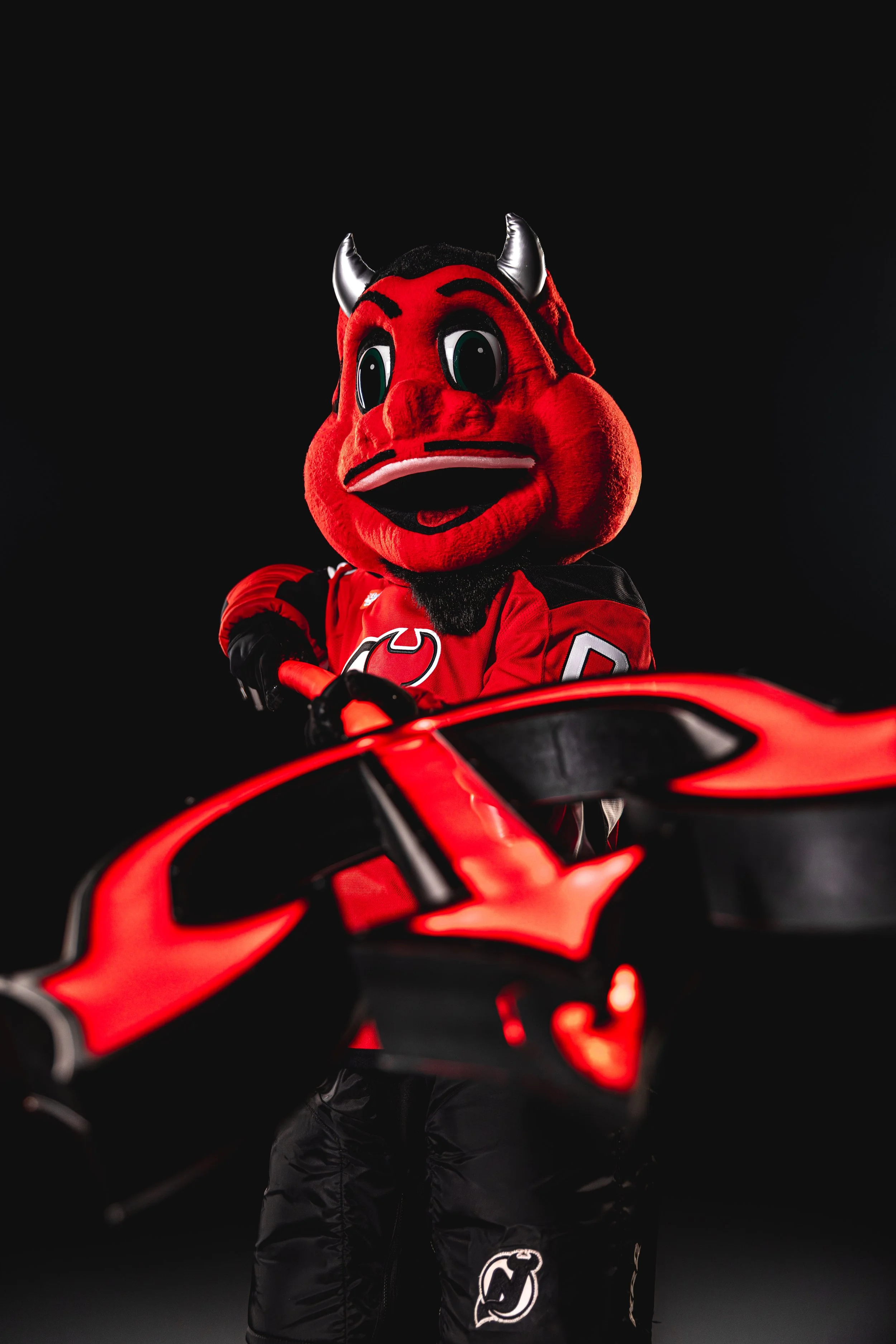 A person dressed in a red devil mascot costume with large eyes, black eyebrows, metallic horns, and a red face. They are wearing a red sports jersey with black and white details, including the logo of the New Jersey Devils ice hockey team, and holdin