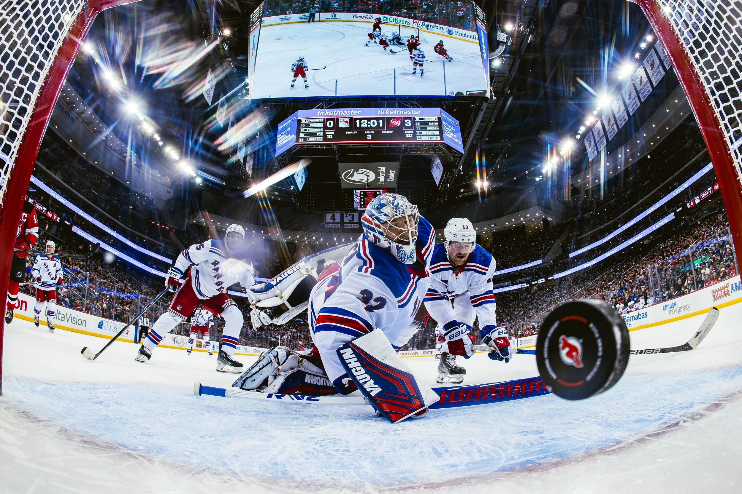 Hockey players on ice rink during a game, with a scoreboard showing 12:01 remaining in the third period, and a large screen displaying the game above. The scene is surrounded by fans in the arena.