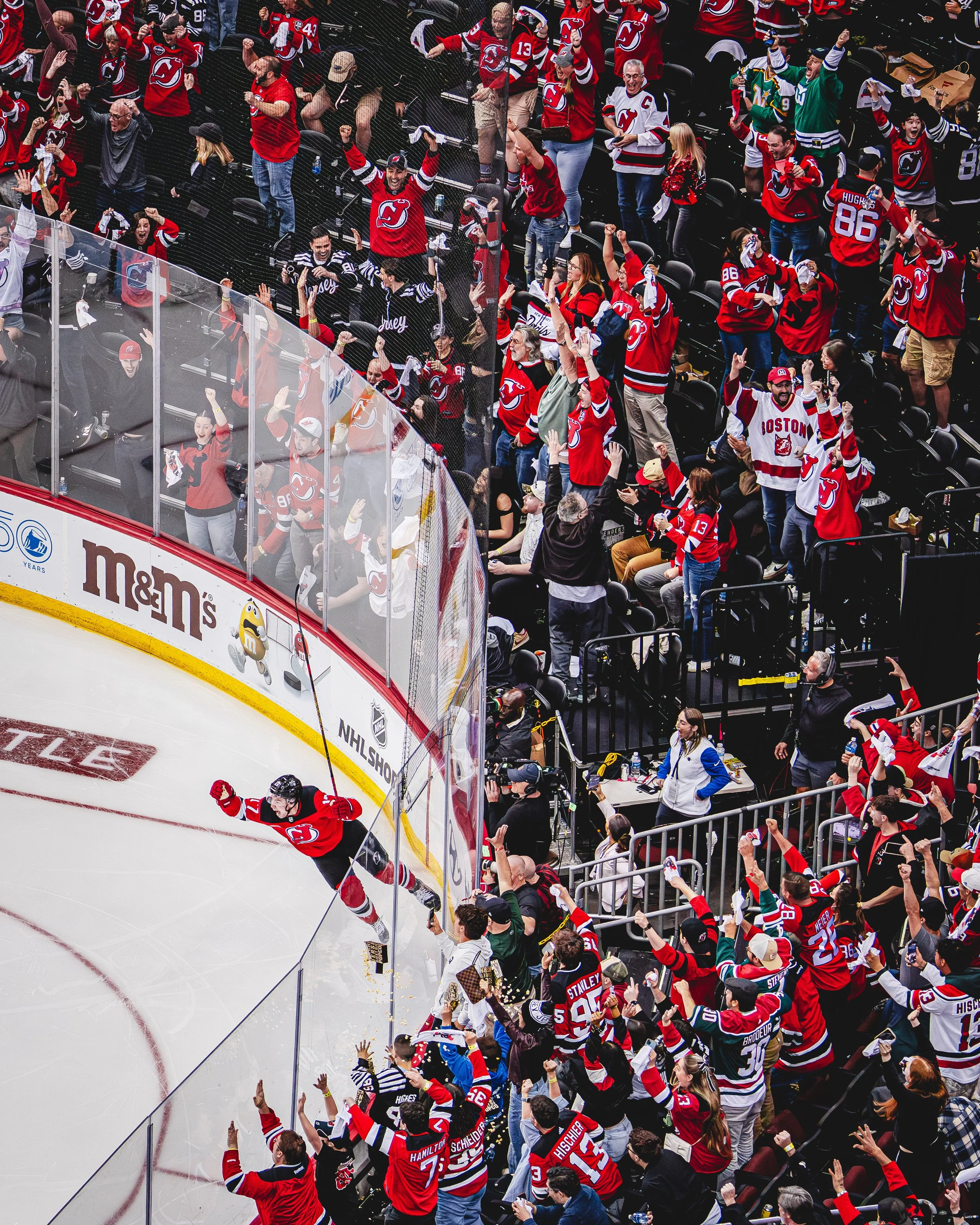 Ice hockey players celebrating a goal near the glass while fans cheer and wave in the stands, with many dressed in red team jerseys.