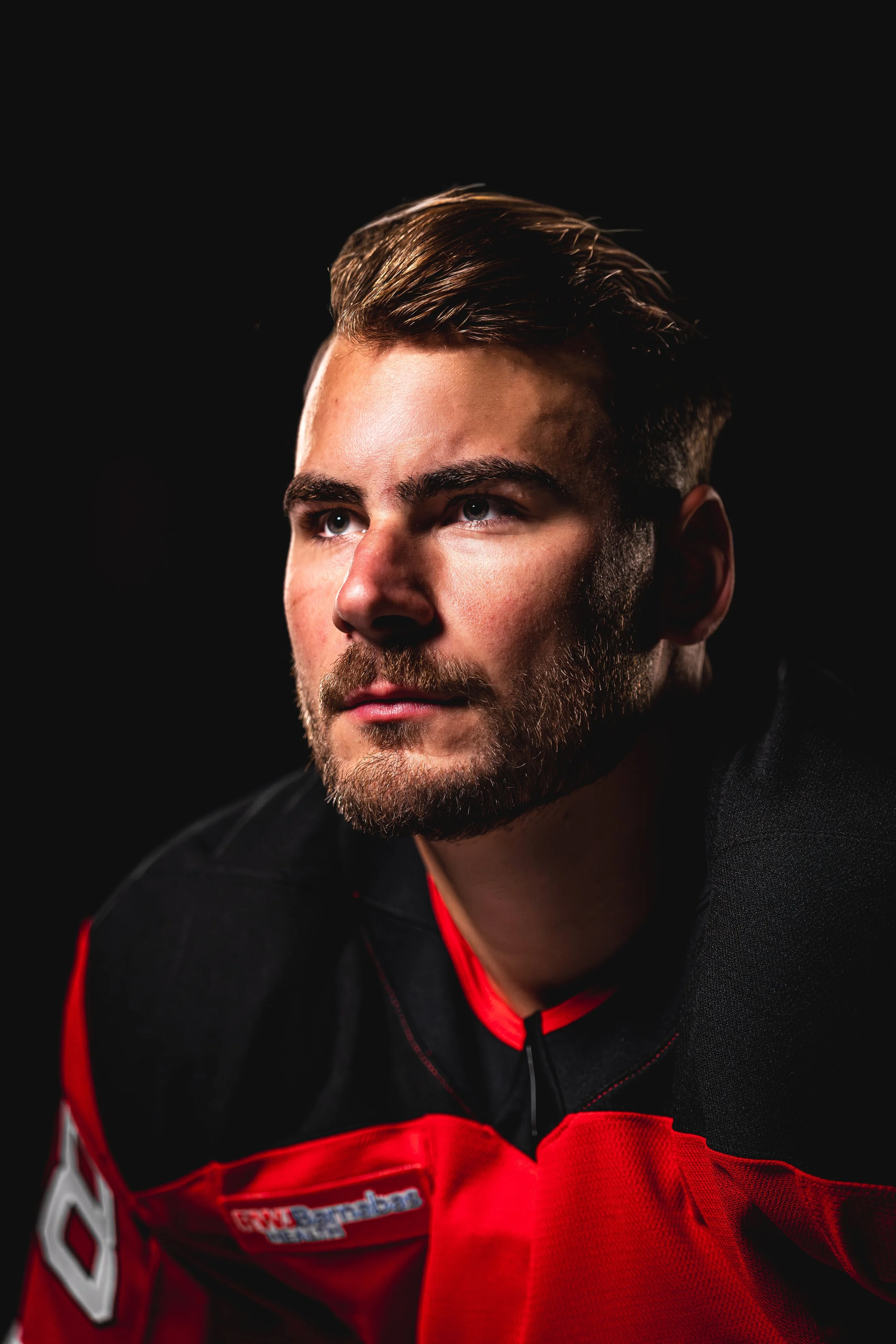 Close-up portrait of a male race car driver wearing a black and red racing suit, looking pensively to the side, set against a black background.