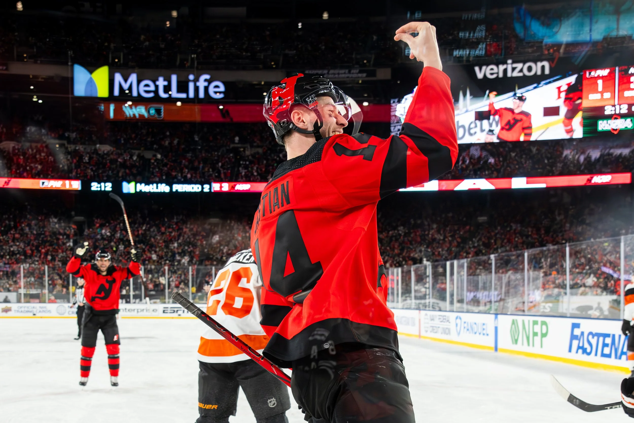 Hockey players on the ice rink celebrating, with one player in focus raising his arm in triumph, while others cheer in the background. There are advertisements and scoreboard visible above in the arena.