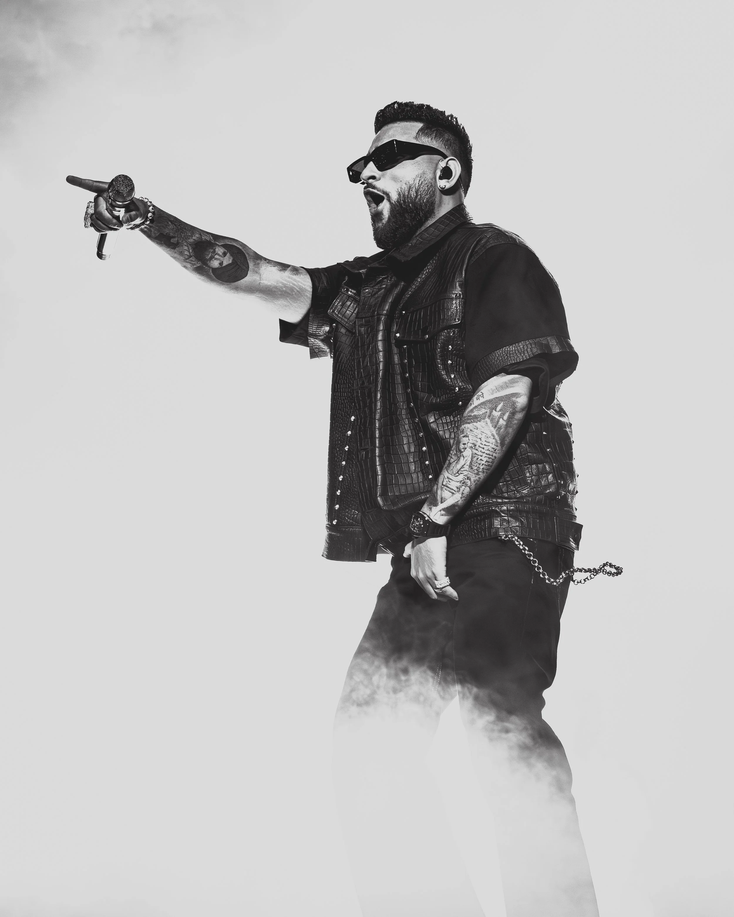 A man with tattoos, wearing sunglasses, a leather vest, and dark pants, singing into a microphone while performing on stage with a cloudy background.