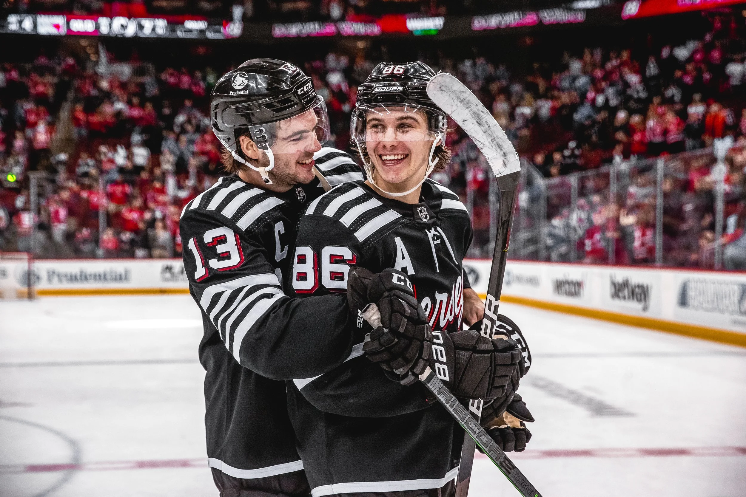 Two ice hockey players in black and white jerseys hugging and smiling during a game, with a crowd in the background.