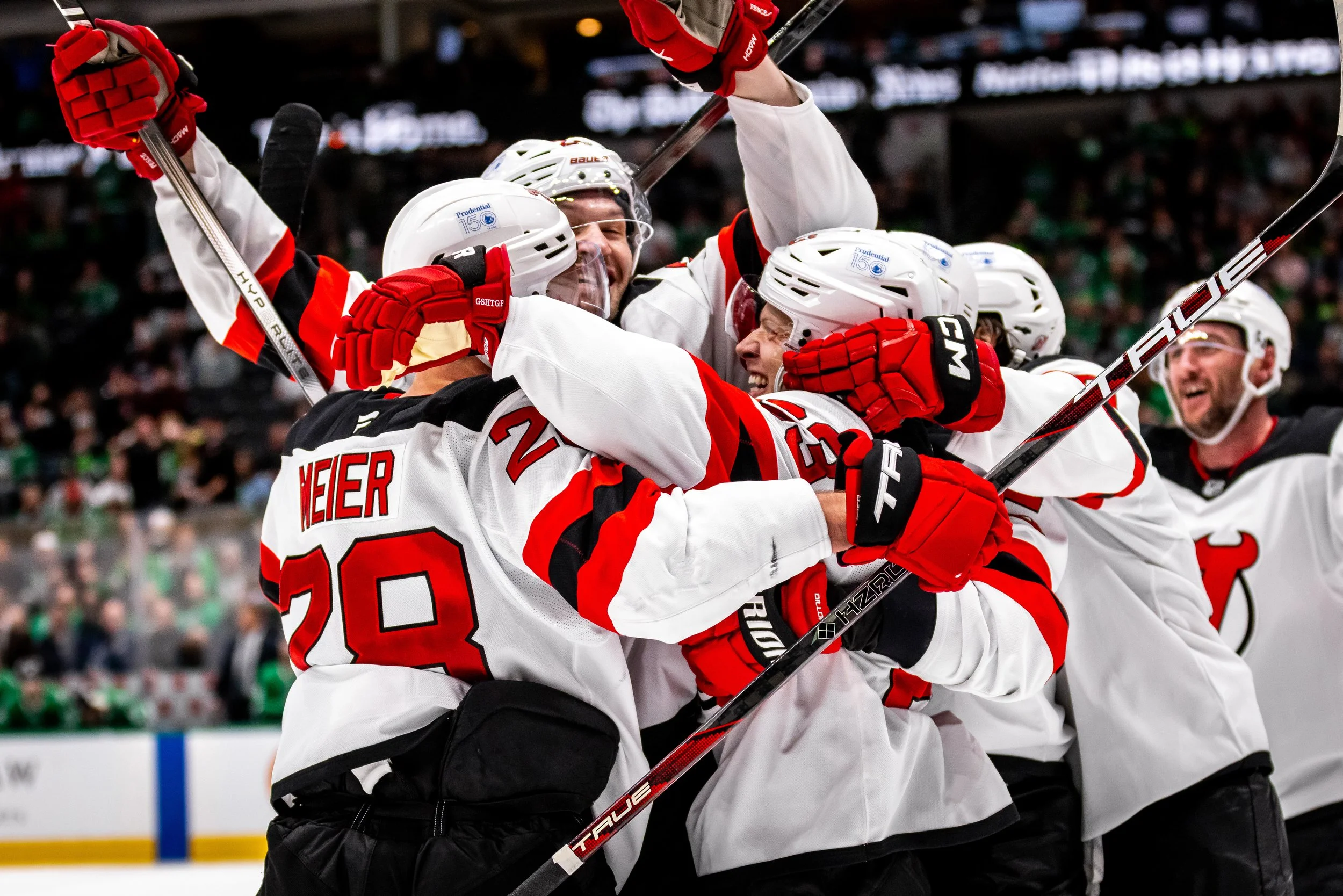 Hockey players in white and black jerseys celebrating on ice during a game, some with arms raised, in an indoor arena with a blurred audience in the background.