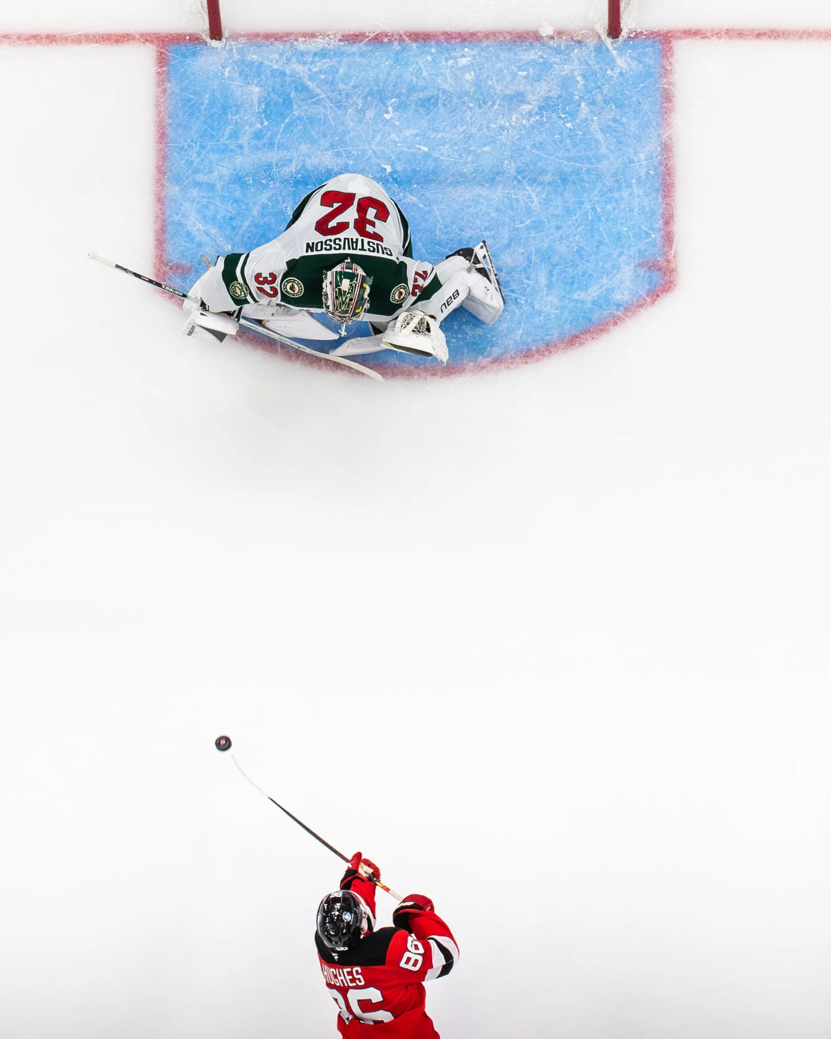 An ice hockey player in a red and black uniform with the number 86 on the back, shooting a puck towards the goal. The goalie is in white and green gear, with the number 32 on the jersey, positioned in front of the net on ice.