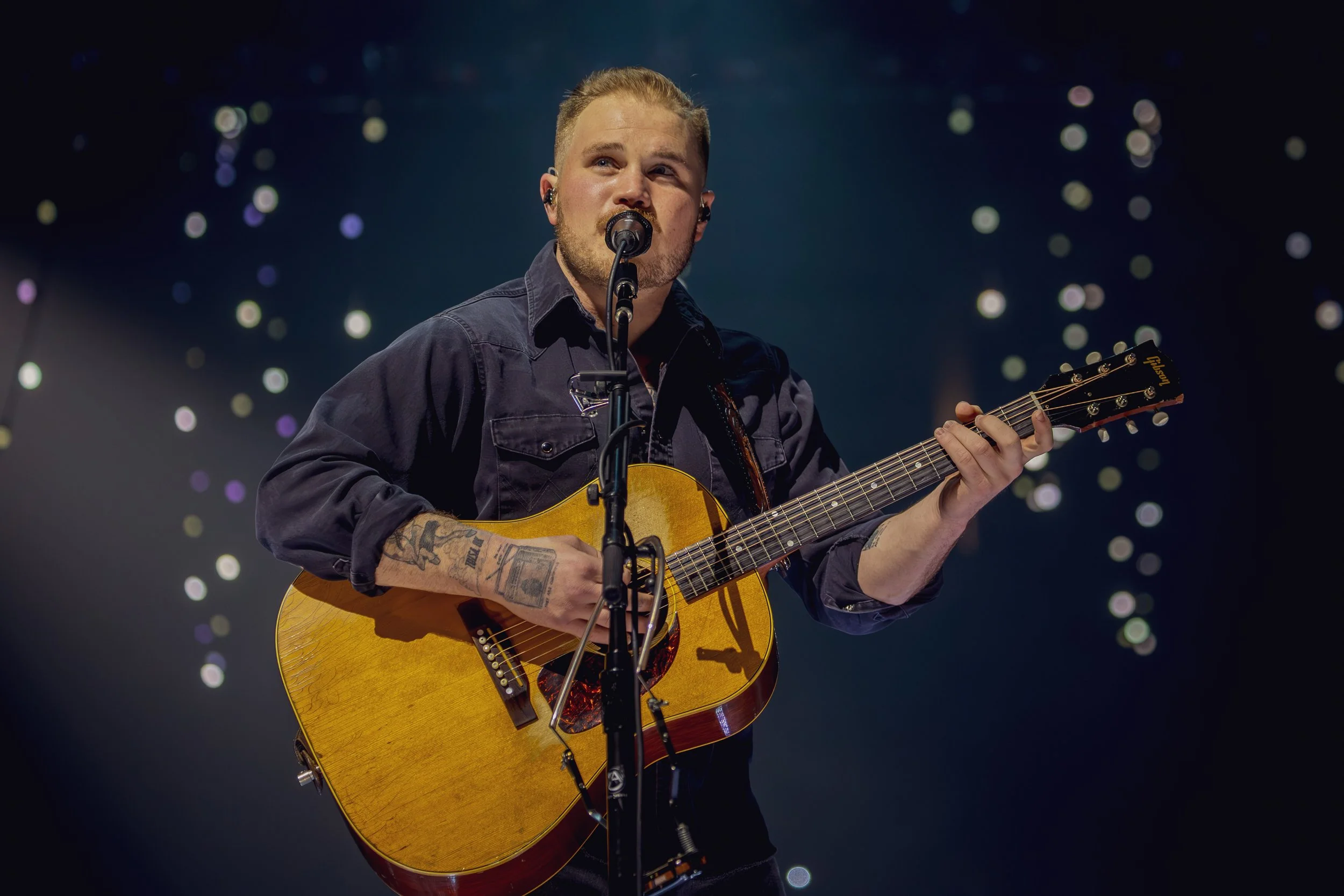 A male musician with tattoos on his left arm playing an acoustic guitar and singing into a microphone on stage with a background of out-of-focus stage lights.