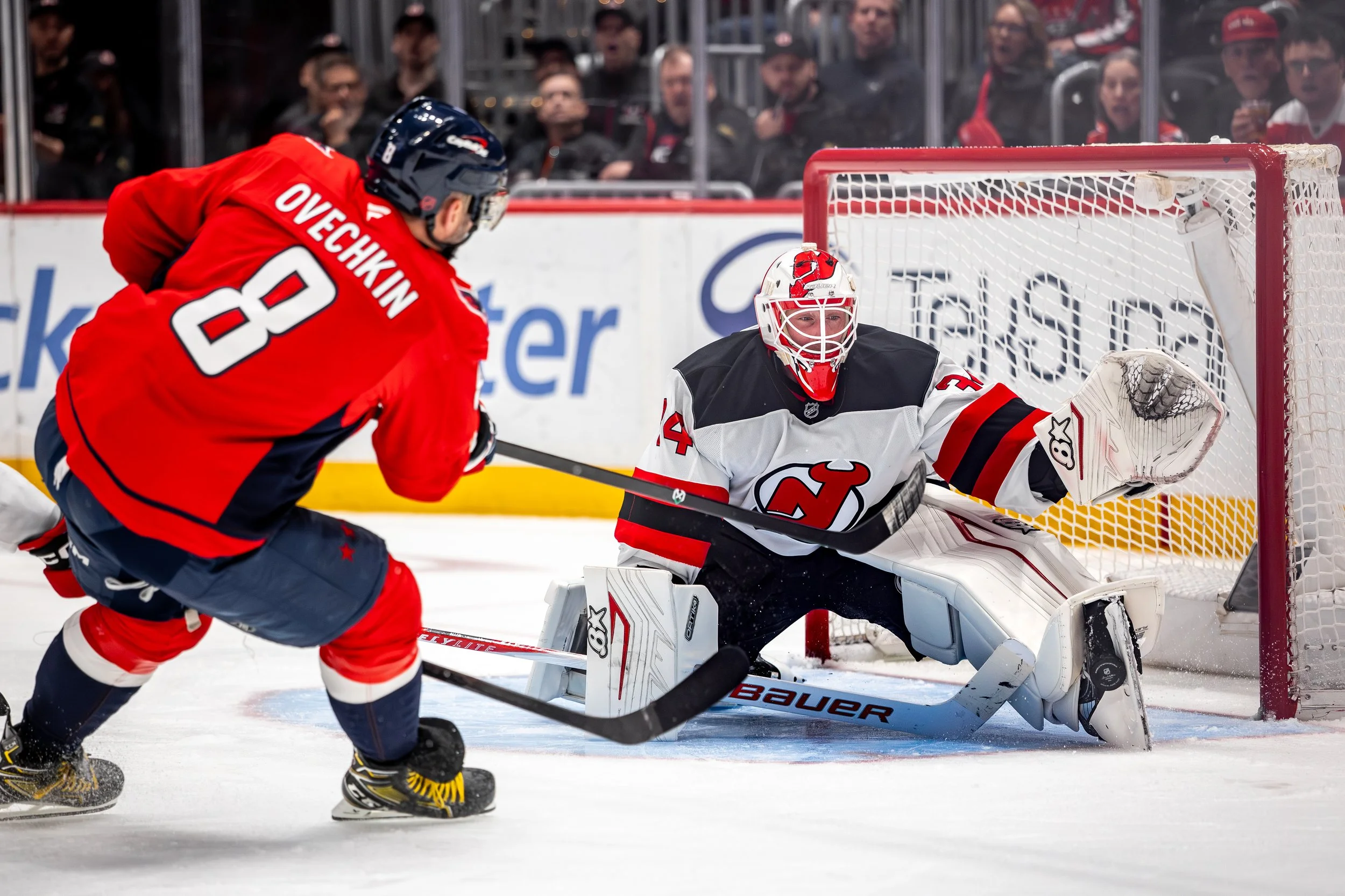 Hockey players in action during a game, with one player shooting the puck towards the goal where the goalie is ready to block it.