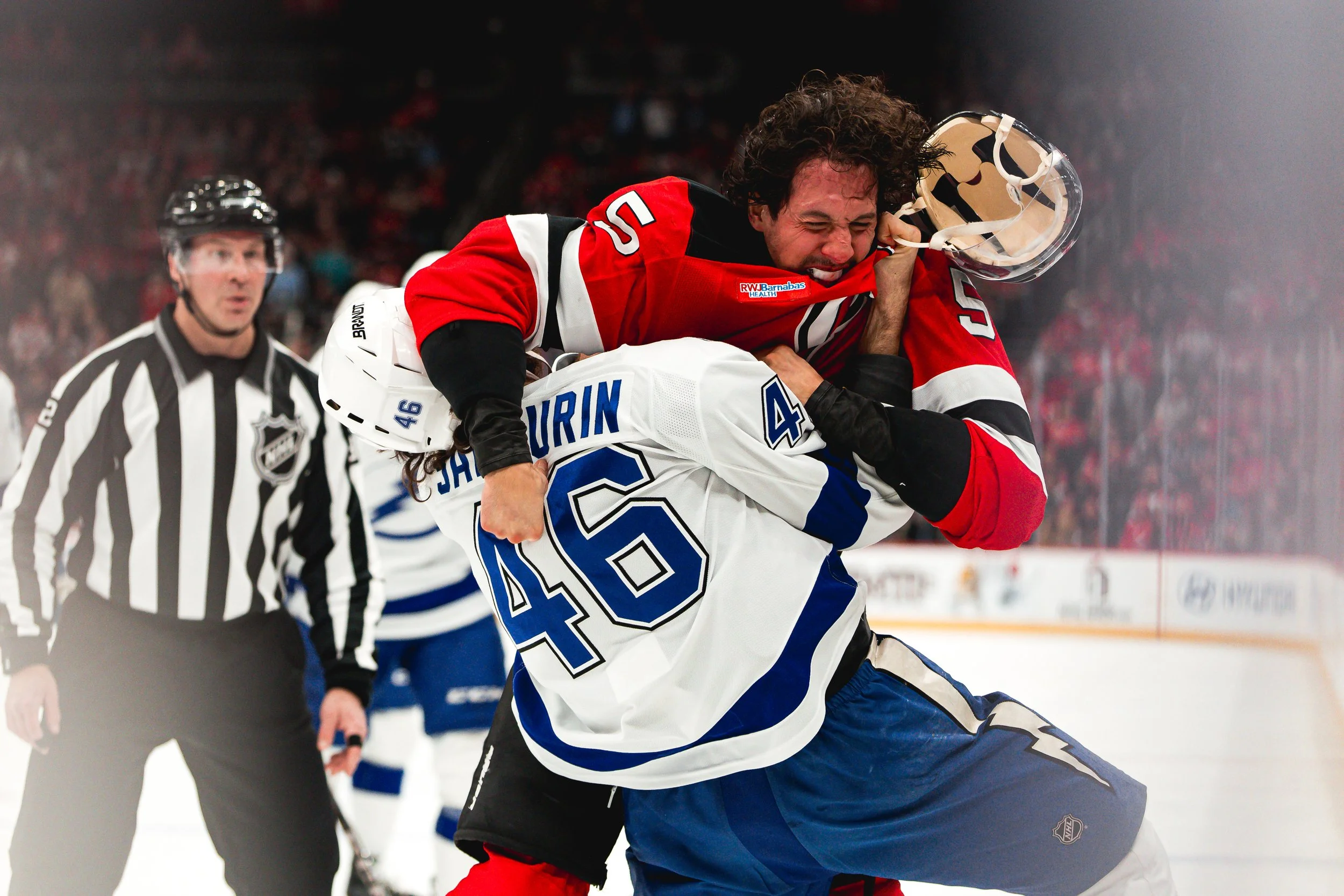 Two hockey players are fighting on the ice during a game. One is from the Toronto Maple Leafs with the number 46, and the other is from the New Jersey Devils with the number 56. A referee in the background watches the altercation.