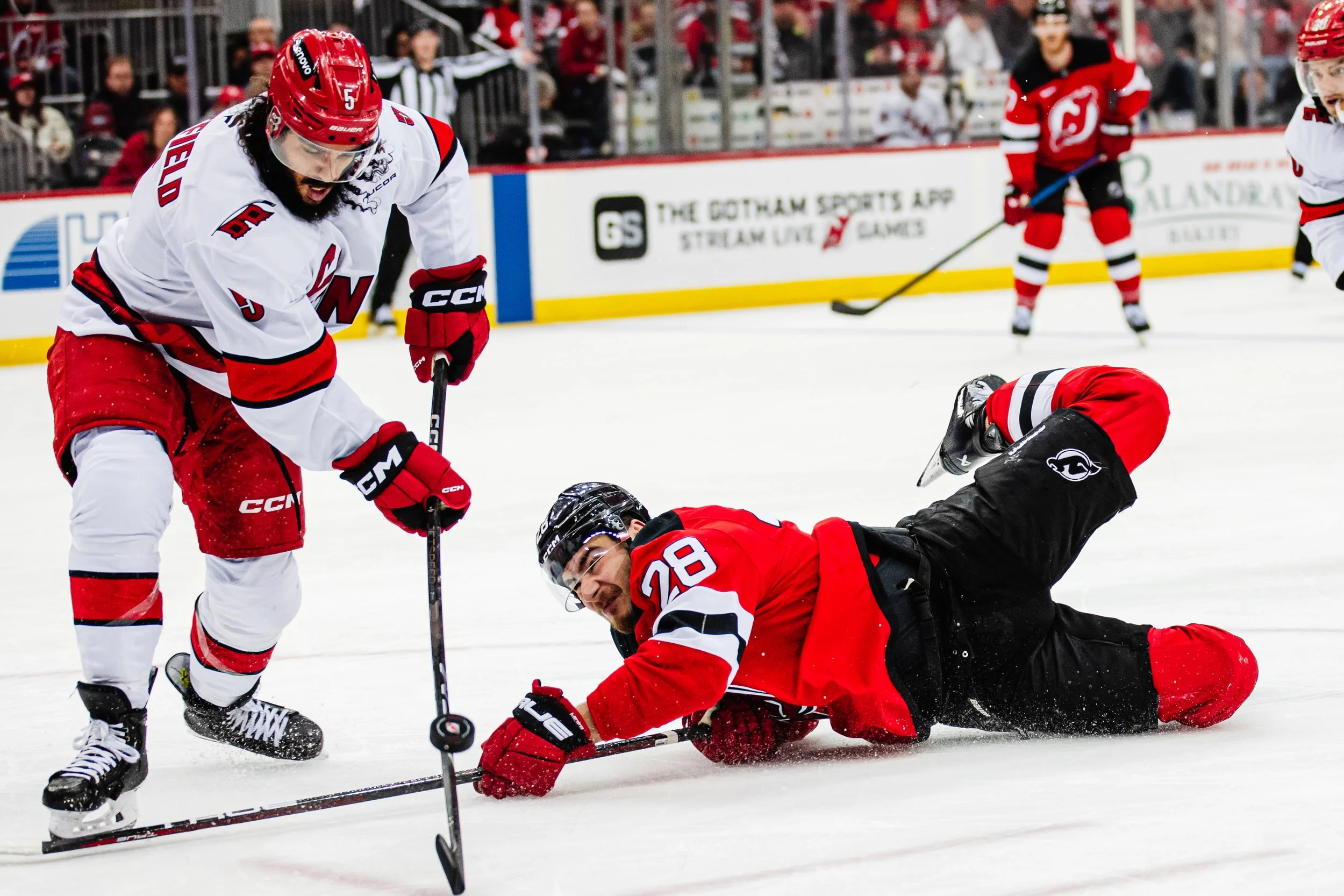 Hockey players on the ice, with one player falling to the ground and others surrounding him, during a game. Spectators are visible in the background.