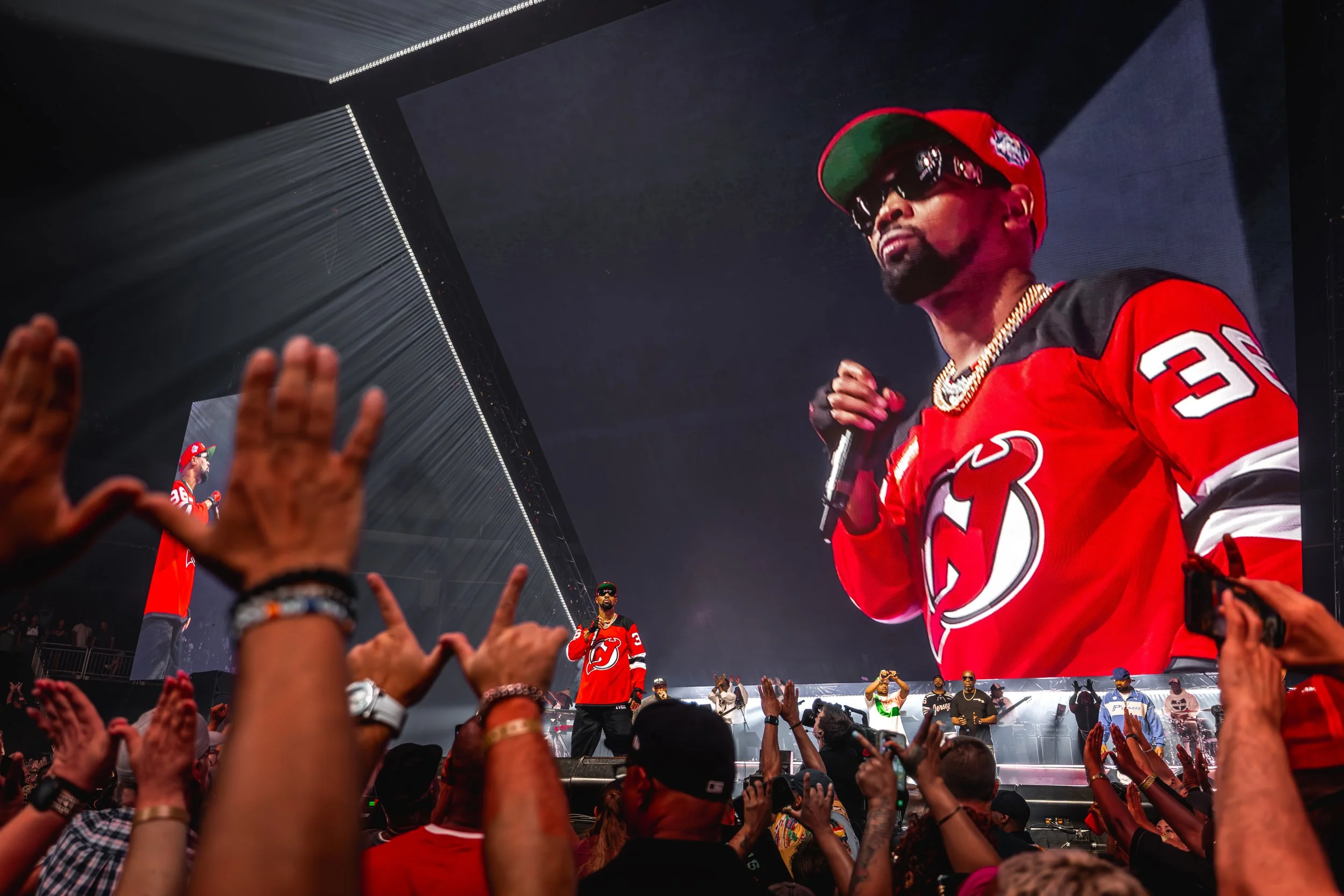 A large screen displaying a man in a red sports jersey and a red cap, holding a microphone, with hands and fans in the foreground.