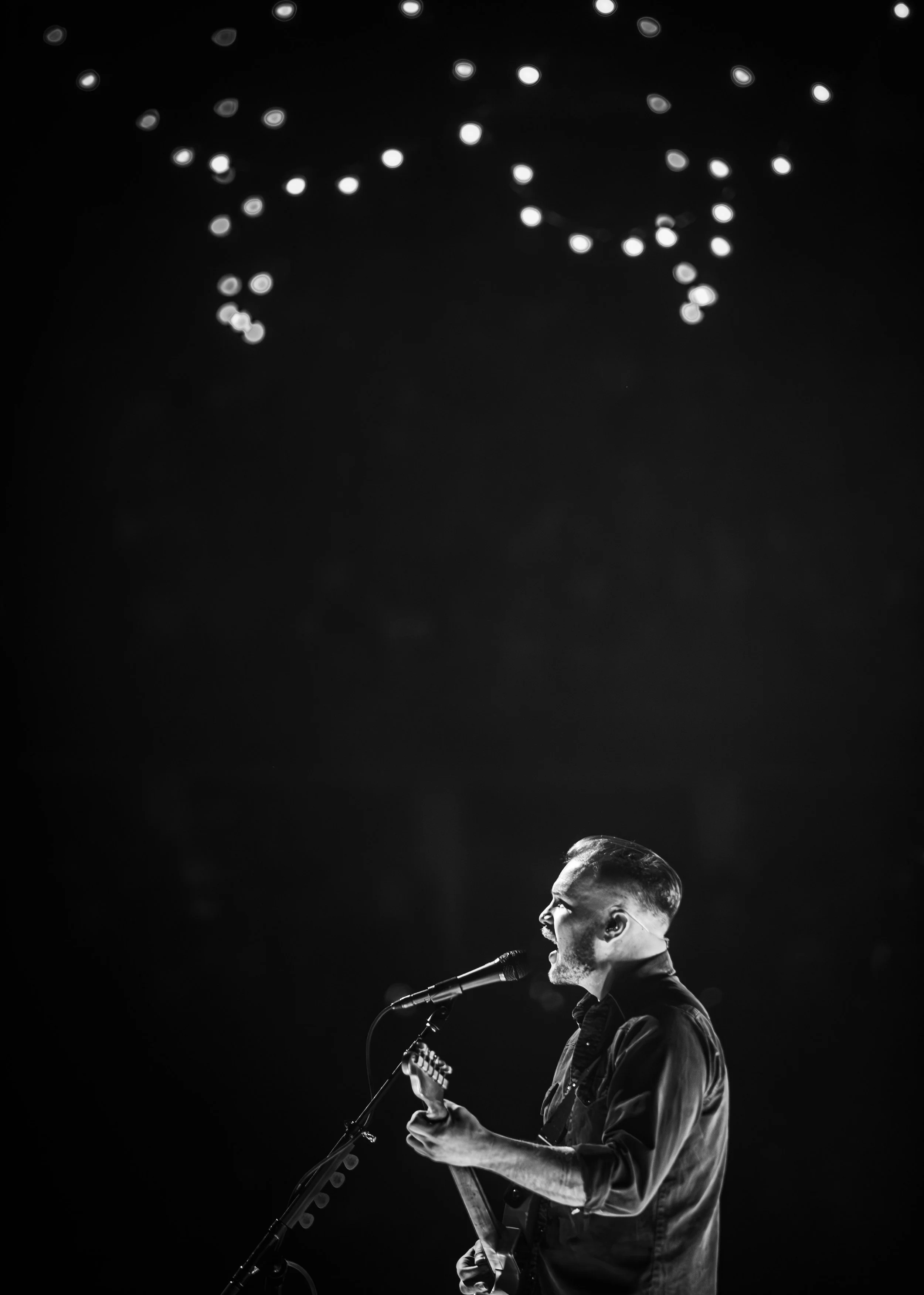 A male musician with a beard singing into a microphone and playing an acoustic guitar on stage, lit by spotlight with a dark background.
