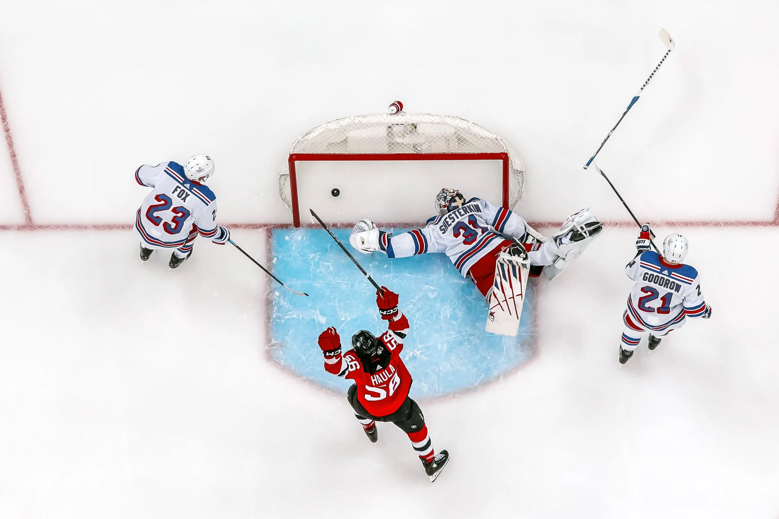 An ice hockey game with players from two teams near the goal. The player in red is reaching for the puck, while the players in white and blue jerseys are defending, with one goalie on the ice attempting to block the shot.