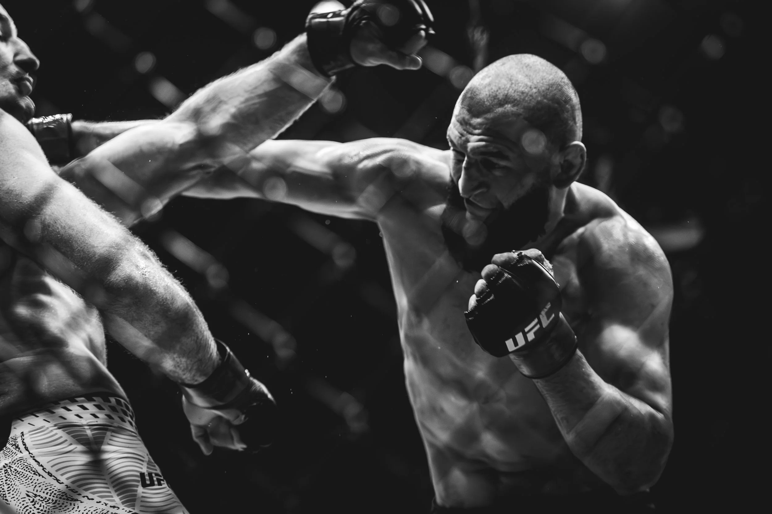 Black and white photo of two mixed martial arts fighters inside a cage, one delivering a punch to the other's face, with a chain-link fence in the background.
