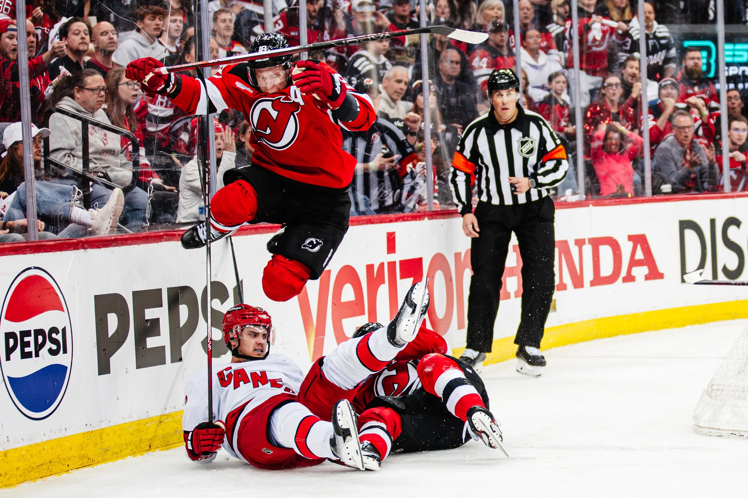 An ice hockey game where one player in a red jersey is celebrating after another player in a white jersey has fallen. The celebrating player is jumping with a hockey stick raised. A referee is watching nearby, and a crowd of spectators is visible beh