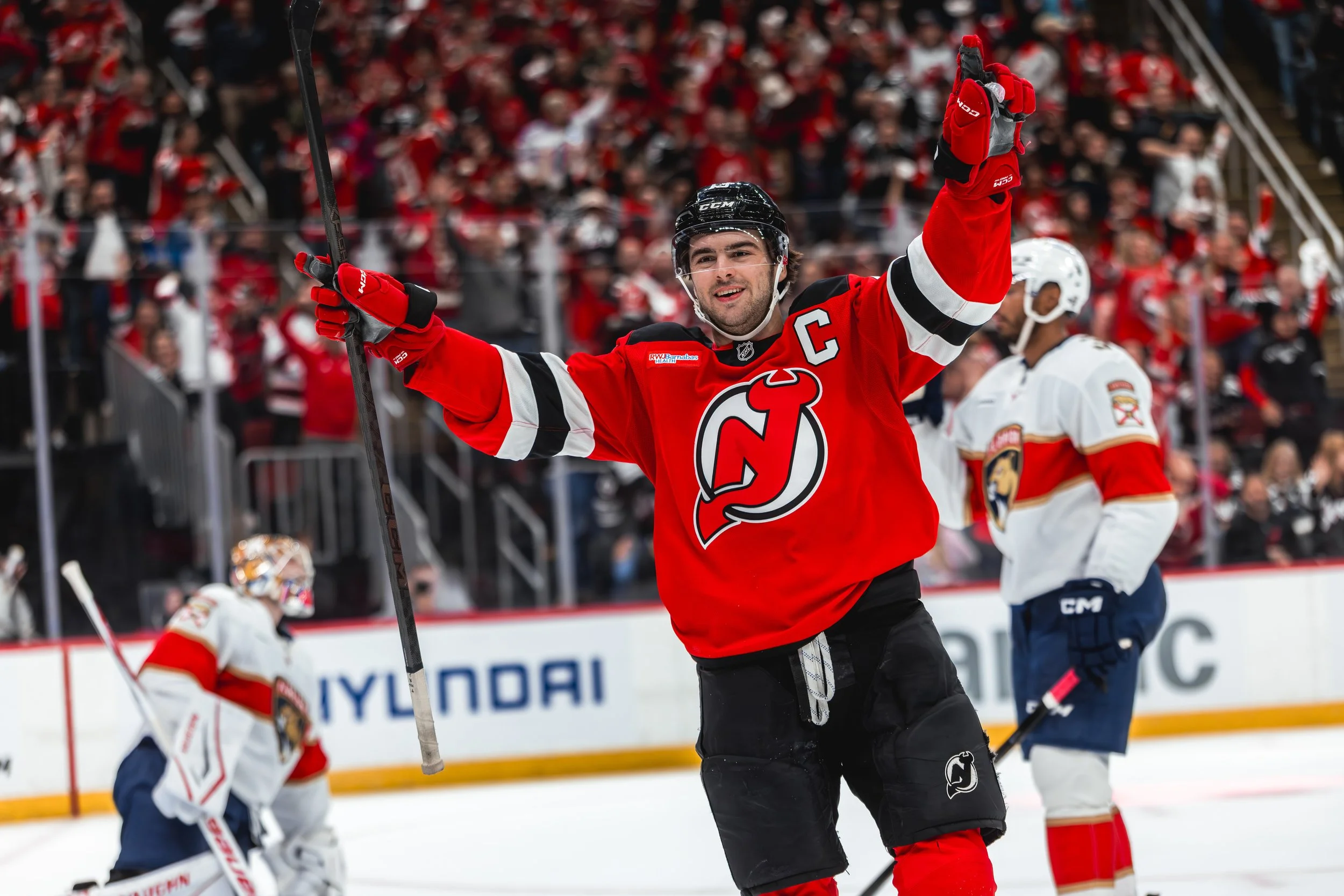 Hockey player celebrating with a raised arm, wearing a red jersey with a black helmet, on an ice rink, with a crowd in the background.