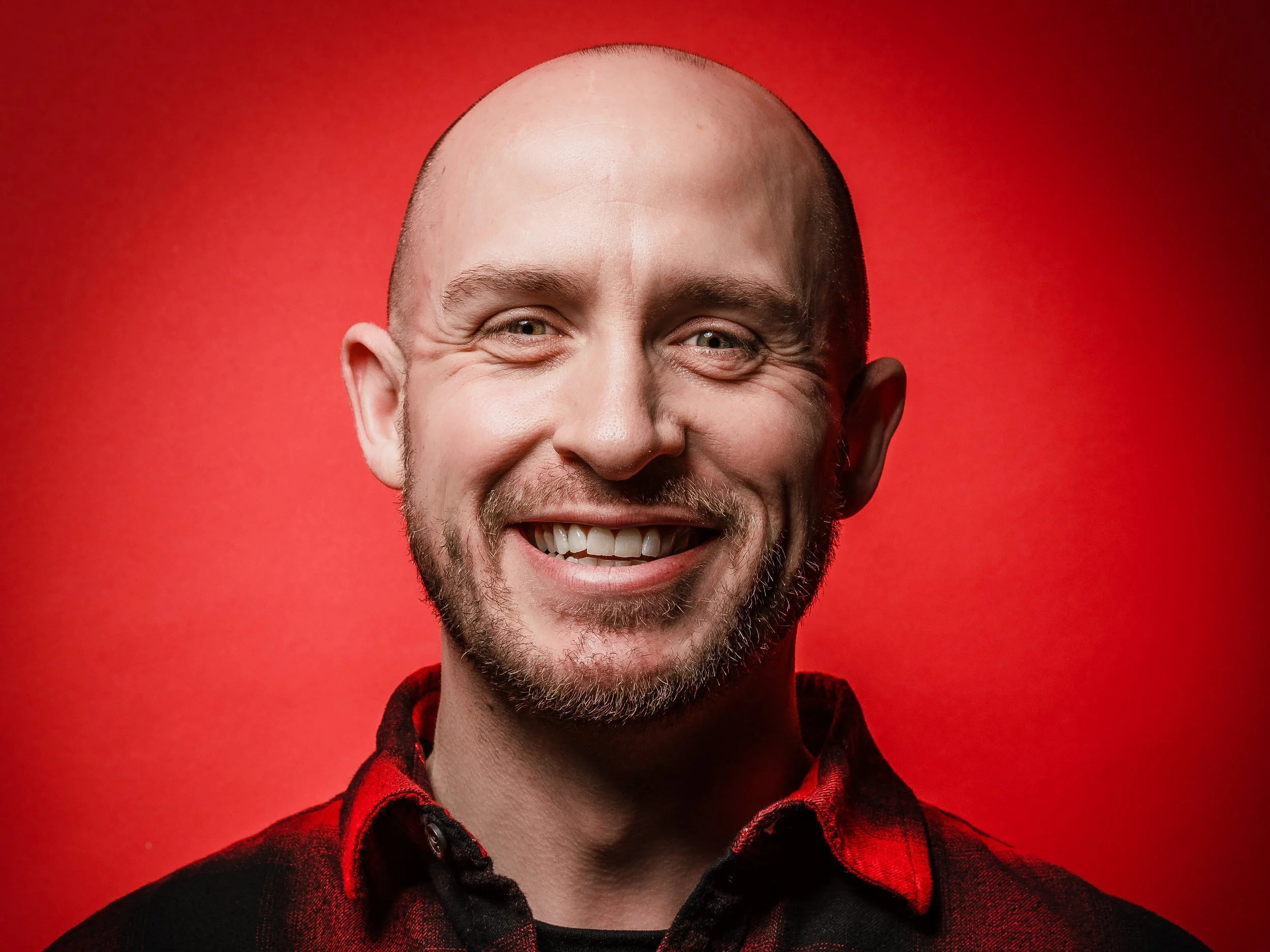 A smiling man with a beard, wearing a red and black flannel shirt, against a red background.