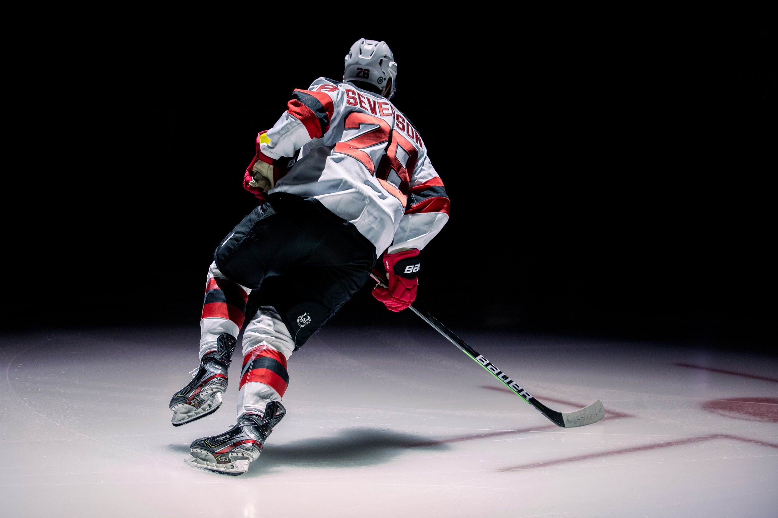 An ice hockey player wearing a white, red, and black uniform with the number 28 and the name 'Seven' on the back, skating on ice with a hockey stick, against a dark background.