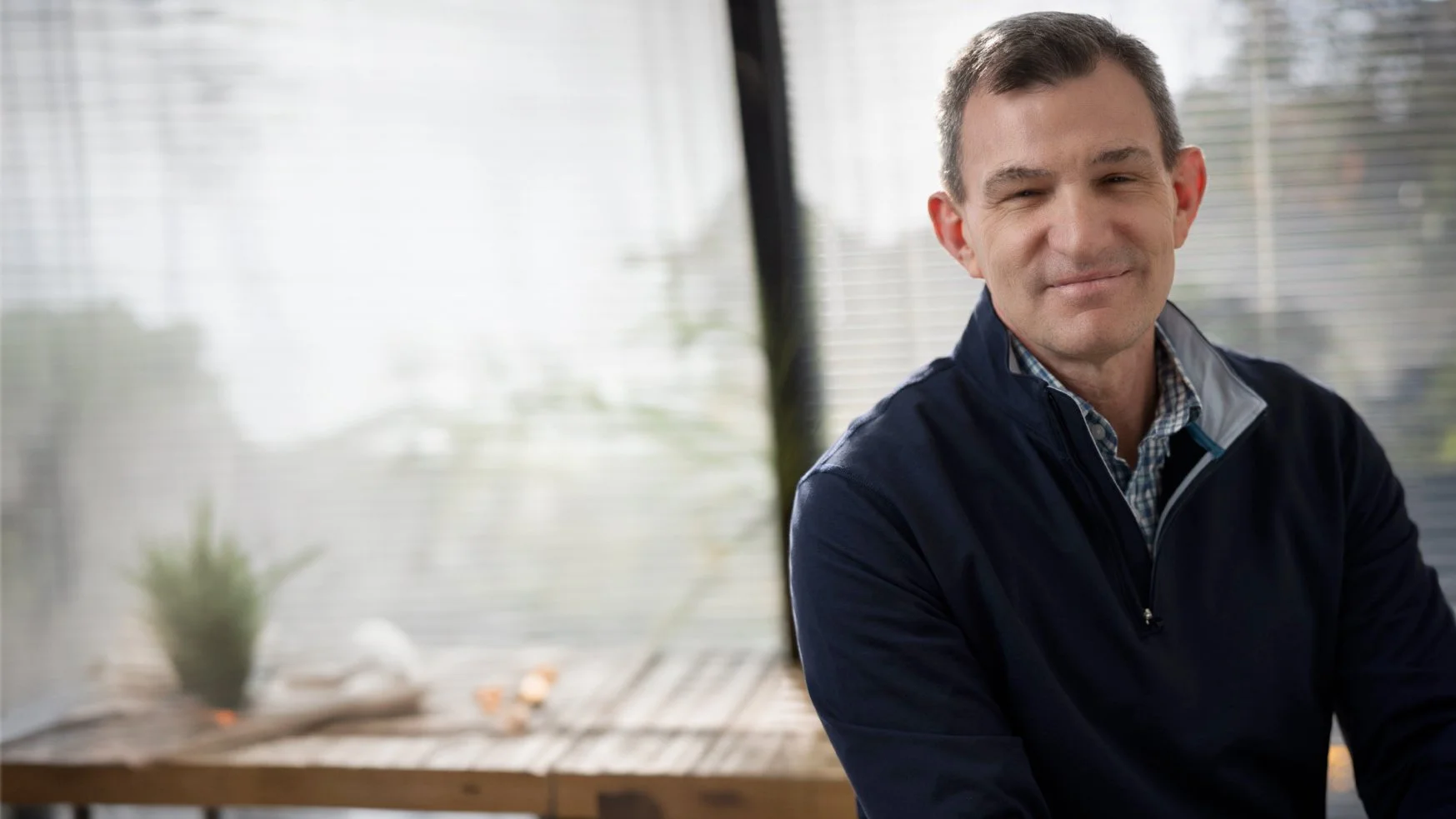 A middle-aged man with short dark hair wearing a navy jacket and checkered shirt, smiling slightly, sitting at a wooden table indoors near a window with blinds.