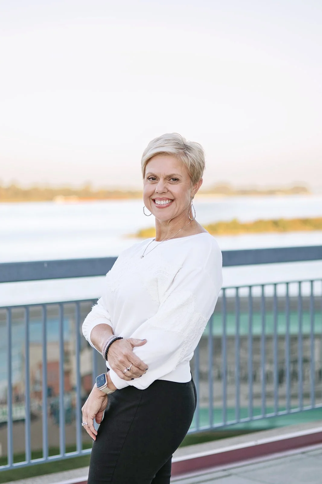 Kim Cates, a certified professional stress and motivational coach, smiling with short blonde hair, wearing a white blouse, hoop earrings, a necklace, and bracelets, standing in front of Paducah, Kentucky riverfront