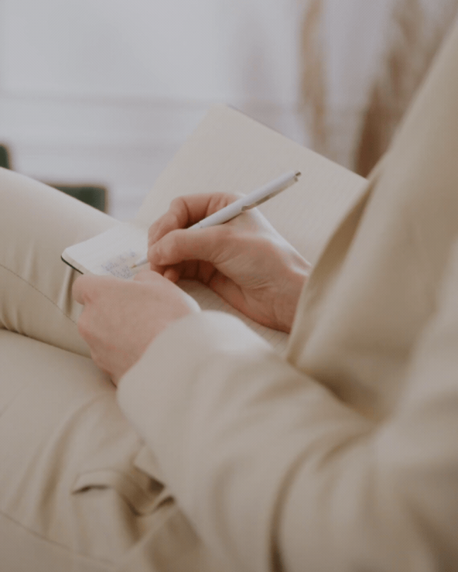 Kim Cates Coaching sitting on a beige couch, writing in a notepad with a pen.