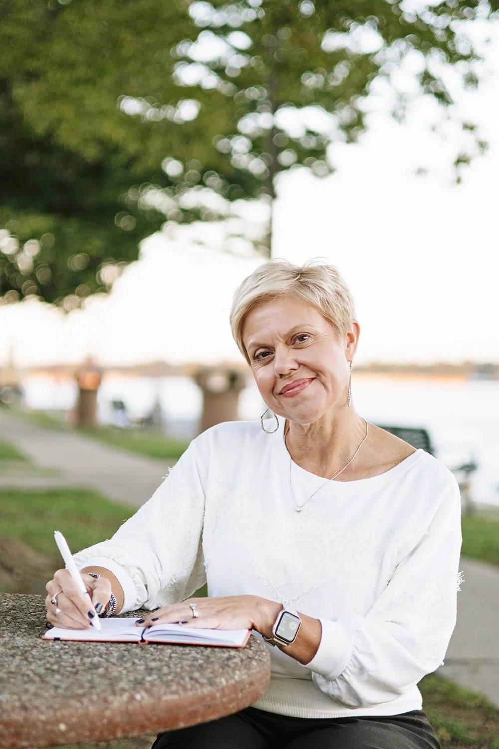 Kim Cates, a certified professional stress and motivational coach, sitting outdoors at a park table near a river, smiling, and writing in a notebook.