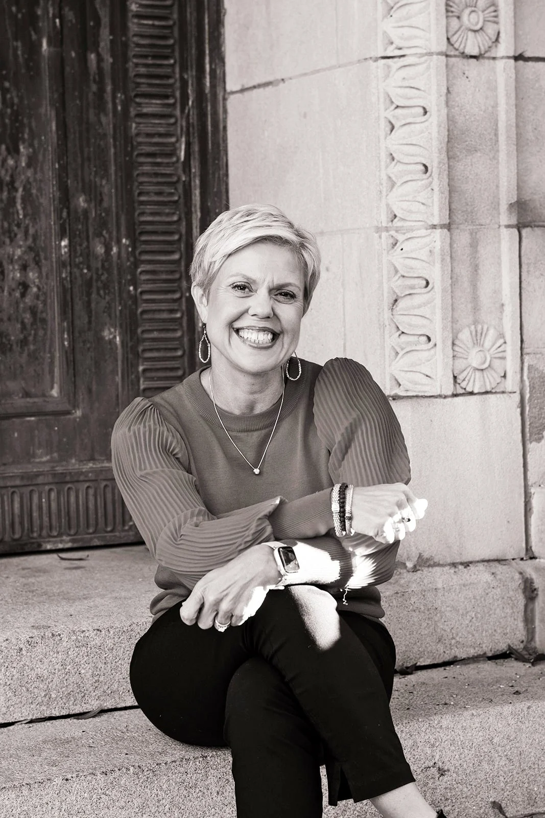 Kim Cates, a certified professional stress and motivational coach, wearing a long-sleeved top, earrings, a necklace, a smartwatch, and bracelets, in front of a decorated stone wall and wooden door.