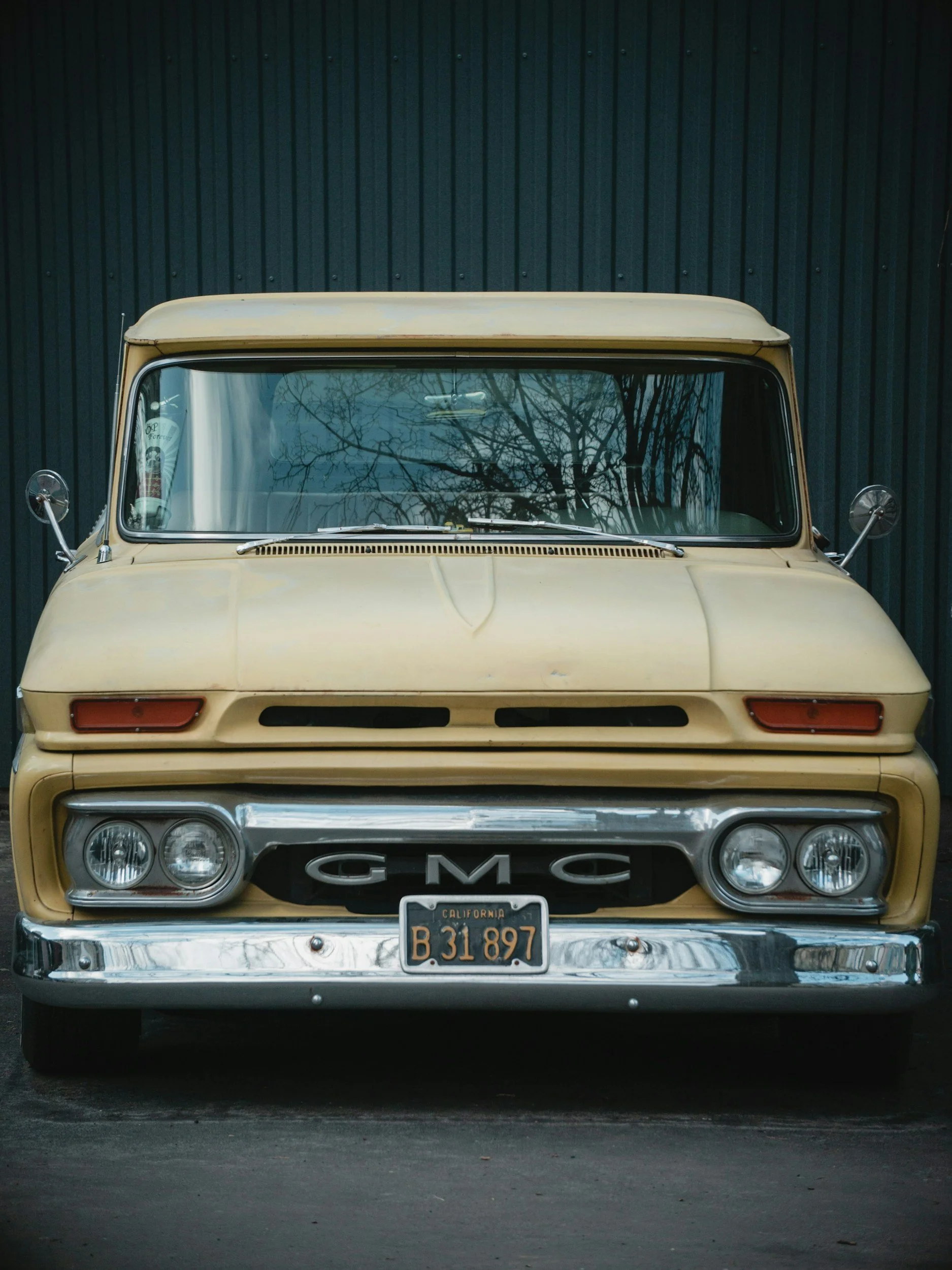 Front view of a vintage beige GMC truck with California license plate B 31 897, parked against a dark vertical-paneled wall.