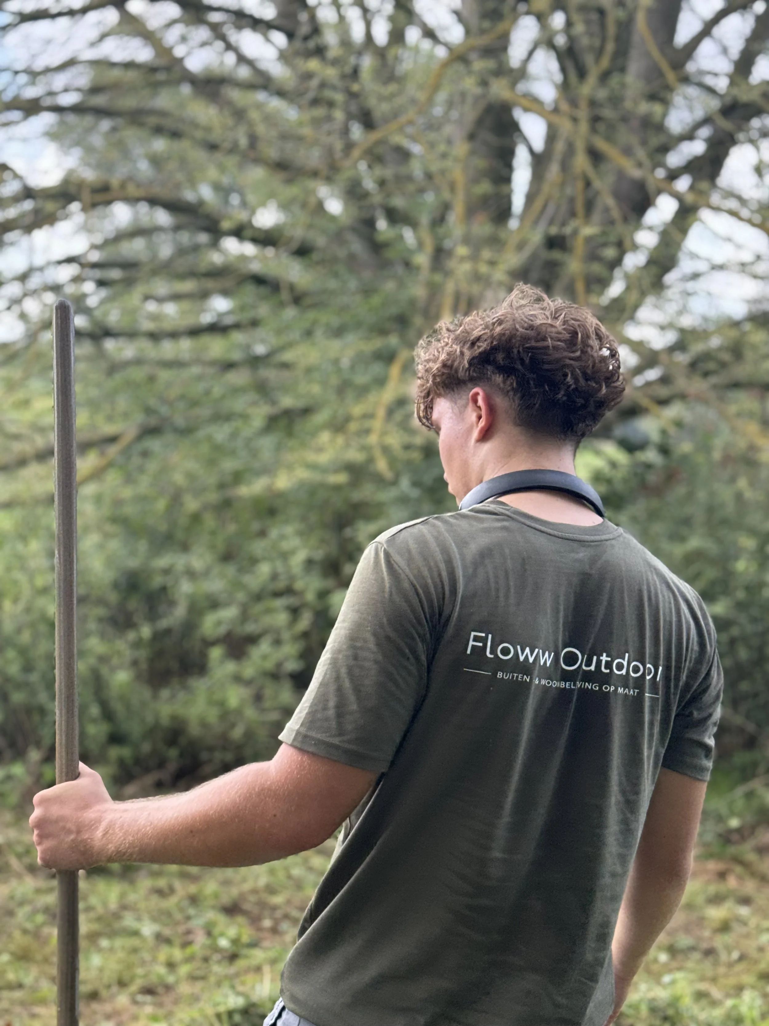 A person with curly hair wearing a dark t-shirt with the text 'Floww Outdoor' printed on it, holding a stick, outdoors in a wooded area with trees and greenery in the background.