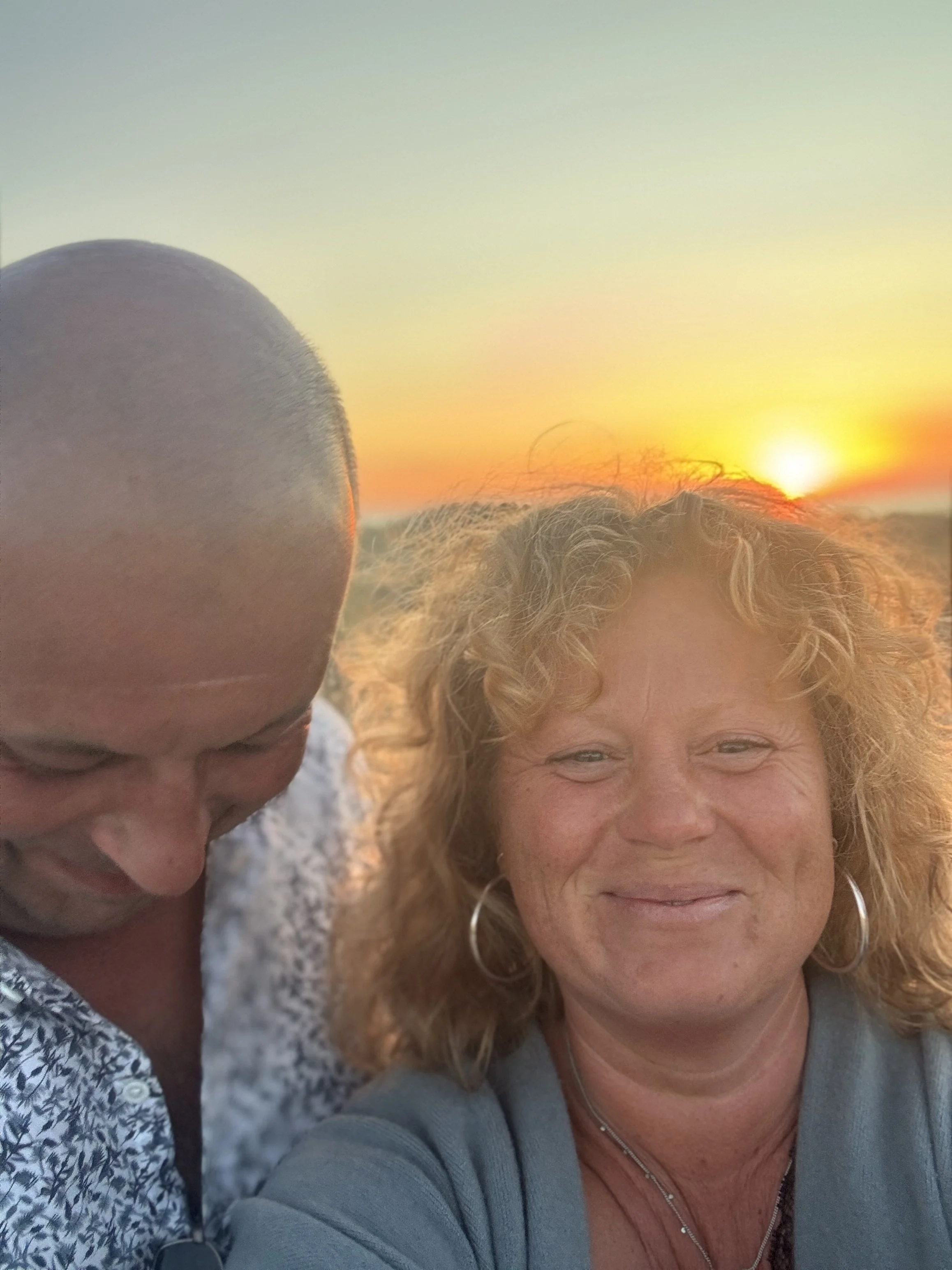 A woman with curly blonde hair smiling at sunset and a man with a bald head looking down, also smiling, outdoors.