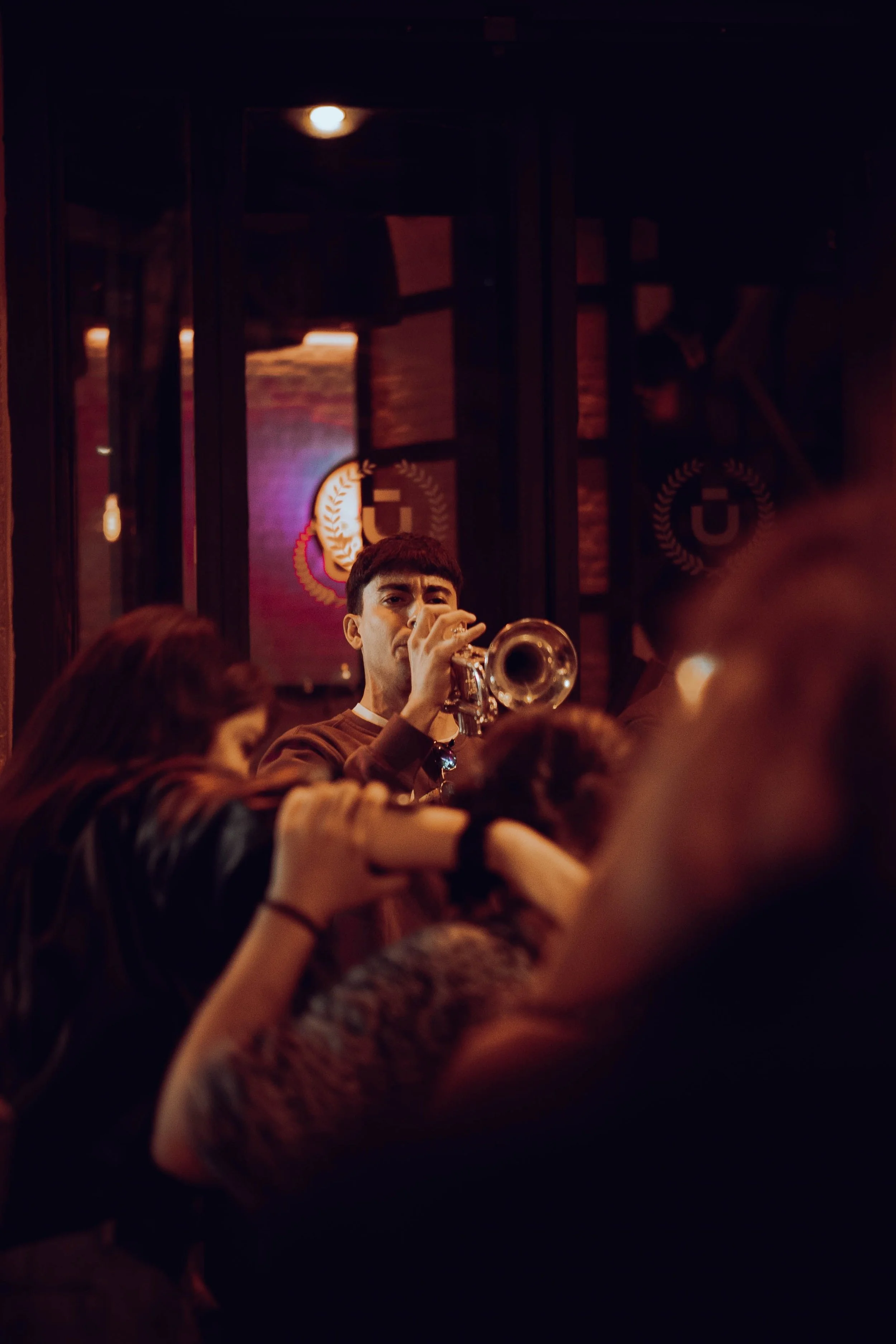 A person playing a trumpet in a dimly lit setting with an audience. The background has dark decor with some illuminated signs or logos.