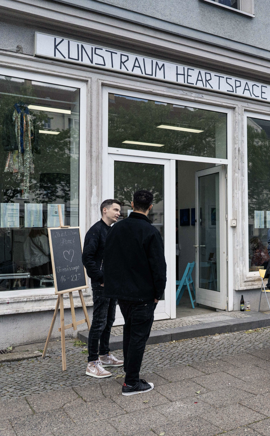 Zwei Männer stehen vor einem Kunstgeschäft namens "KUNSTRAUM HEARTSPACE" und sprechen miteinander. Es gibt ein Holzständer mit einer Tafel, auf der geschrieben steht: "Wo ist dein Herz? Herzreise 16-22°". Der Laden hat große Fenster, durch die Kunstwerke im Inneren sichtbar sind.