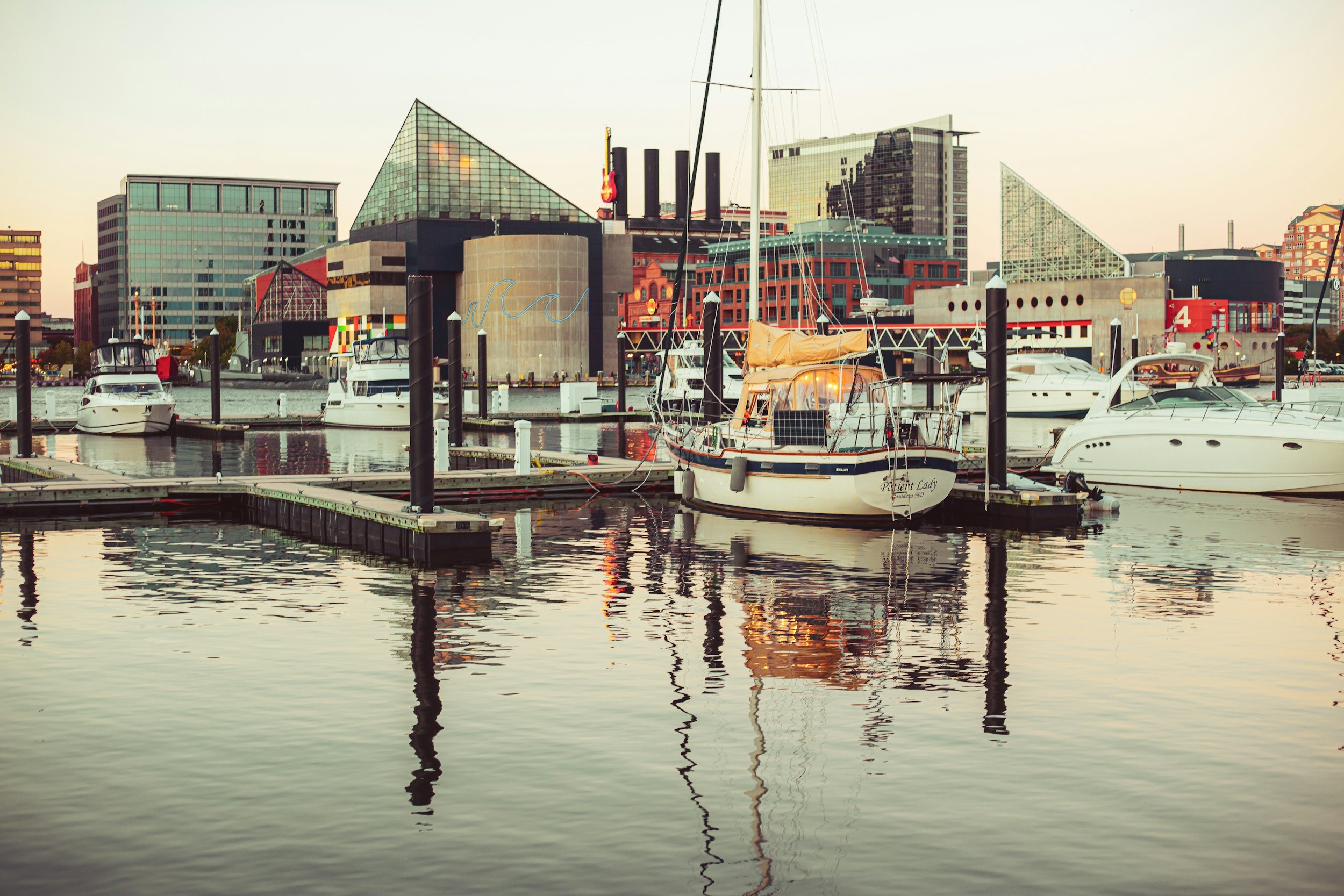 Harbor with boats docked, city skyline with modern buildings in the background at sunset.
