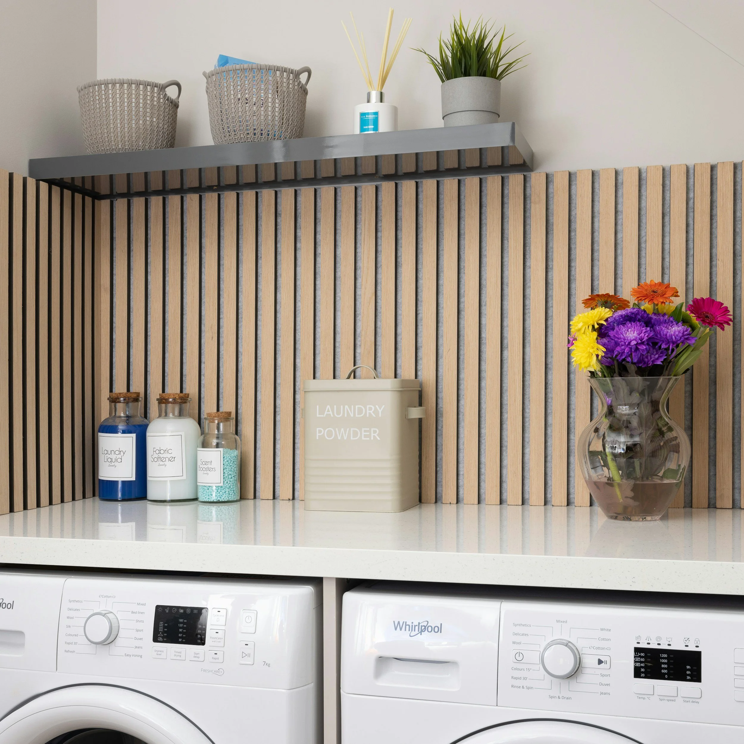 A laundry room shelving unit with storage baskets, a reed diffuser, a potted plant, a laundry powder container, and a colorful bouquet of flowers. Below the shelves are two white Whirlpool washing machines.