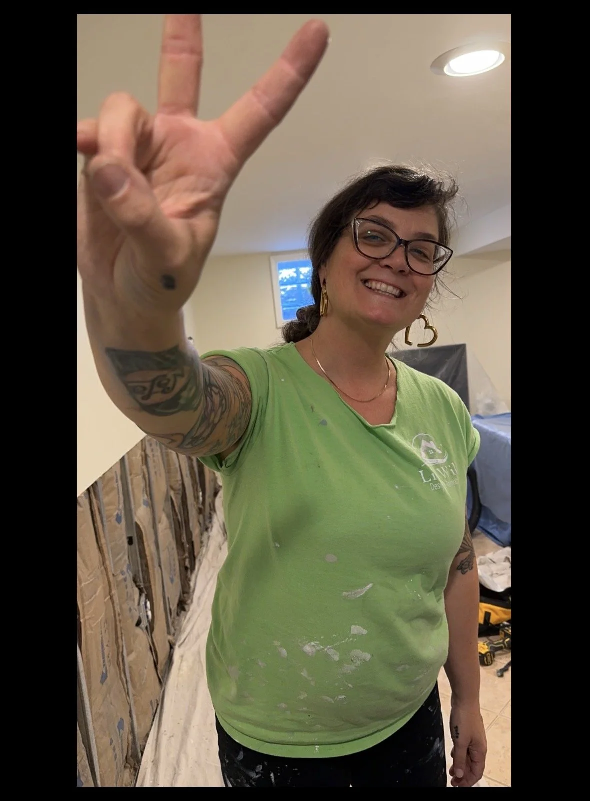 A smiling woman with short dark hair and glasses, making a peace sign with her right hand, wearing a bright green T-shirt with paint stains, standing in a construction or renovation setting with visible drywall and a window in the background.