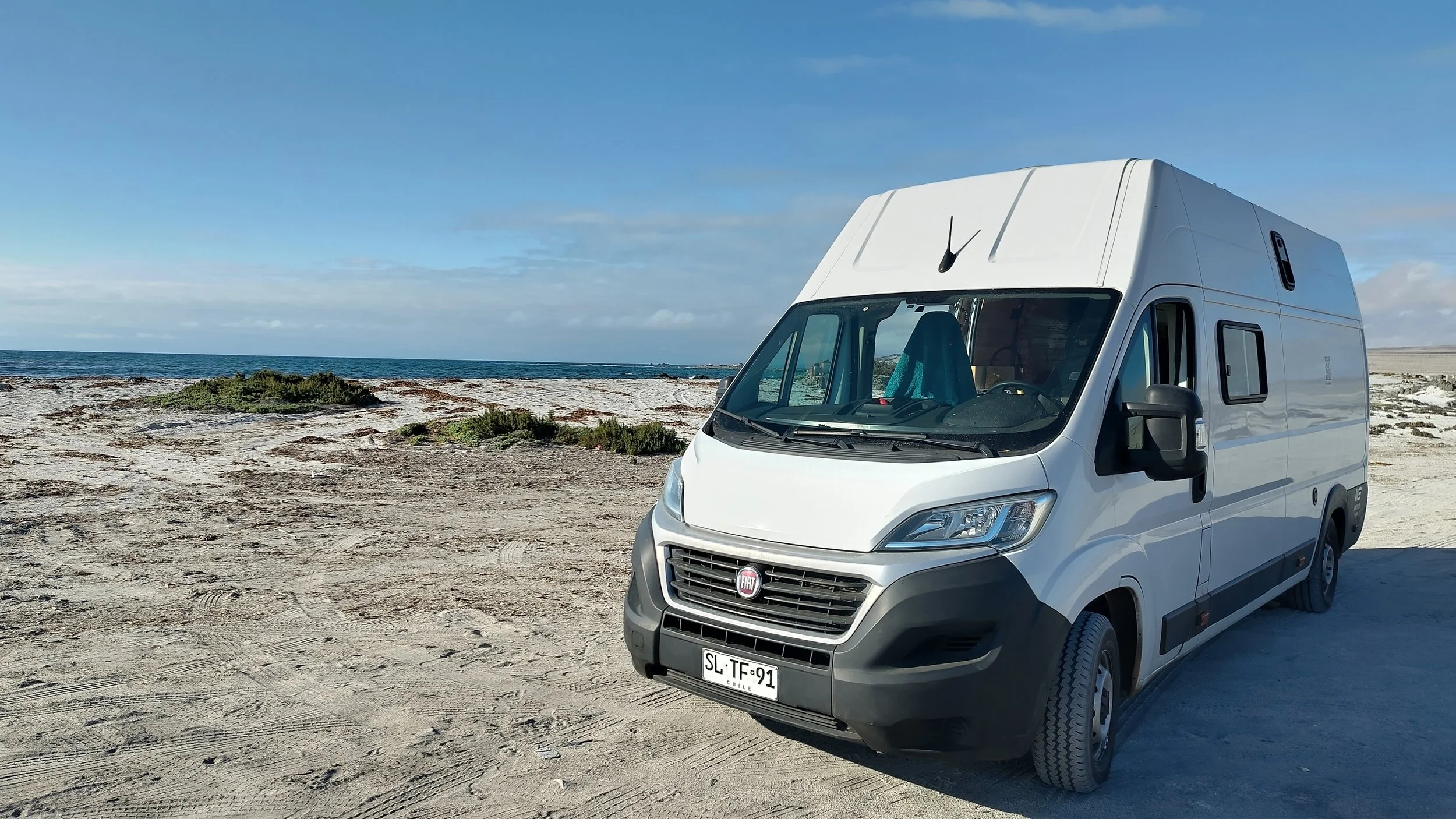 Weißer Wohnmobil steht auf einer sandigen, unbefestigten Fläche am Meer unter einem blauen Himmel mit wenigen Wolken.