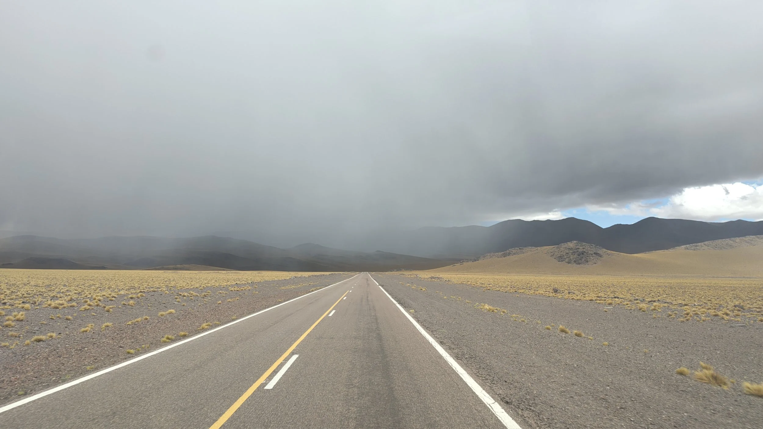 Eine zweiteilige Straße in einer Wüsten-Landschaft mit Bergen im Hintergrund, dunklen Wolken am Himmel und trockenem Gelände mit kleinen Pflanzen
